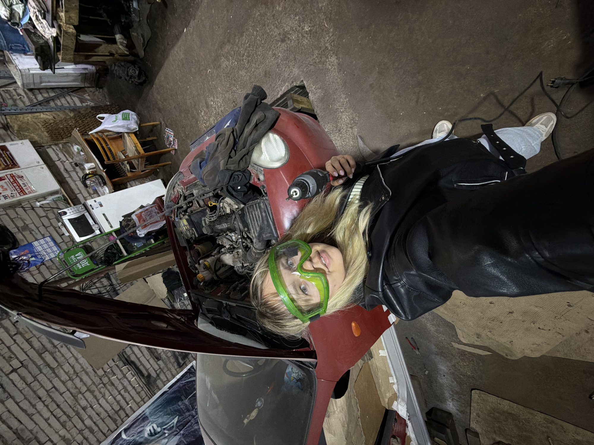 A young, blonde woman is depicted lying on her back on the floor of what appears to be a cluttered garage or workshop, situated in Chaikovsky, Russia. She wears a black leather jacket, light blue jeans, and white sneakers, with bright green protective goggles covering her eyes. In her left hand, she holds a black and silver cordless drill or electric screwdriver, her nails painted light pink.
Next to her, the front section of a red car is visible with its hood open, exposing the engine compartment. Various car components, tools, and rags are scattered around the engine bay. The surrounding environment is typical of a busy workspace, featuring shelves laden with items, cardboard boxes, a wooden chair, and household appliances like a small refrigerator or oven. A white plastic bag with a green logo is visible on the chair. The floor is concrete, and a brick wall can be seen on the left side of the frame.
The woman is engaged in a car repair or maintenance task, indicated by her position under the open hood, the protective goggles, and the tool in her hand. She looks directly at the camera with a neutral expression. The scene is lit by artificial light, suggesting it's either nighttime or a poorly lit indoor area.
Visible text includes a partial "J-PAK" logo on the white plastic bag and some indistinct Russian text on a red poster or sign in the background, along with the number "2010" on another item.