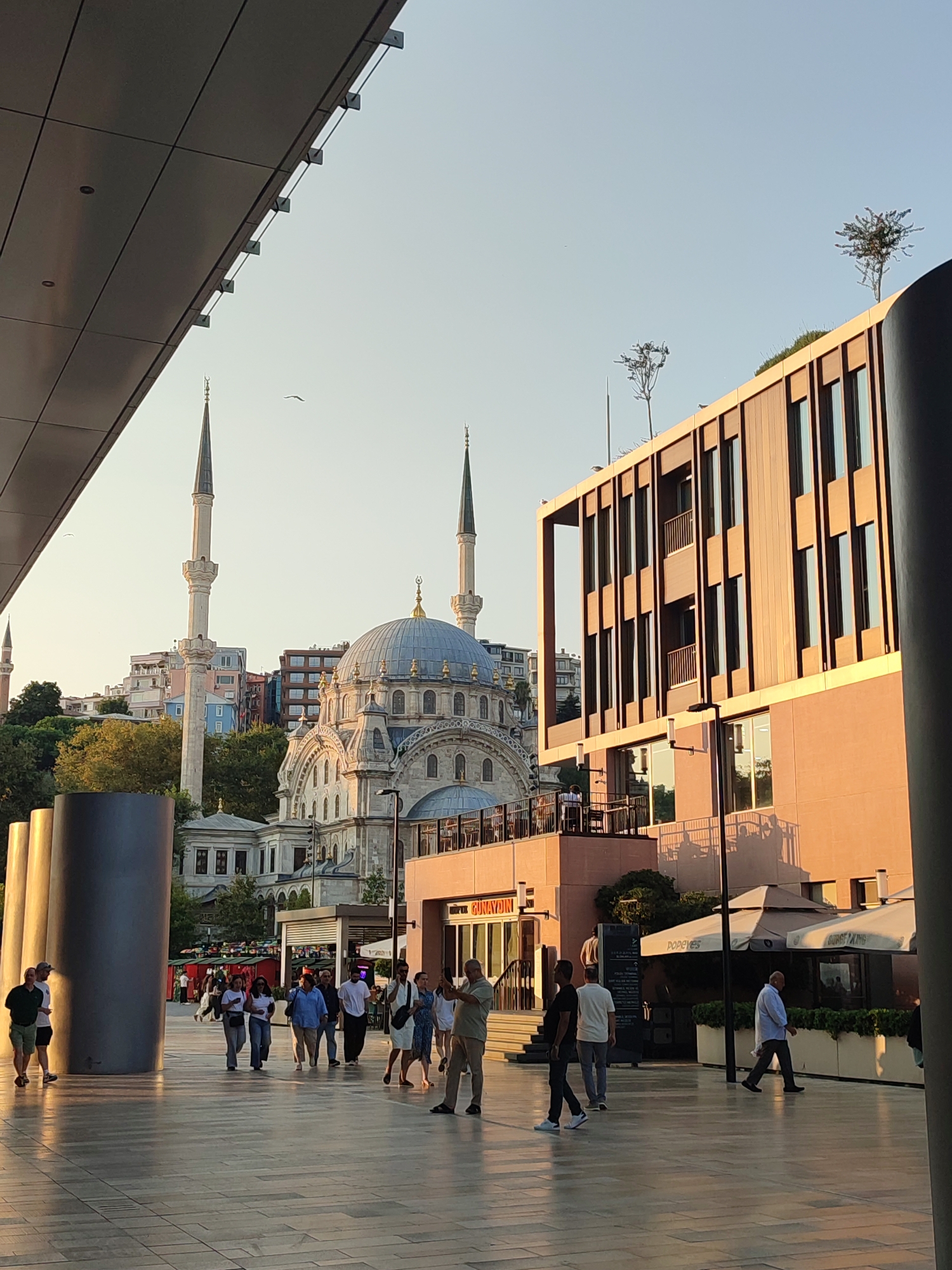 The image depicts a street scene in Beyoğlu, Turkey, with a prominent mosque in the background. In the foreground, several people are walking on a paved walkway. A modern building with a wooden facade and numerous windows stands to the right. To the left, part of a large, dark structure partially obscures the view. In the middle ground, an establishment with outdoor seating and umbrellas is visible. The mosque features a large dome and two minarets. The sky is clear and the lighting suggests it is late afternoon.