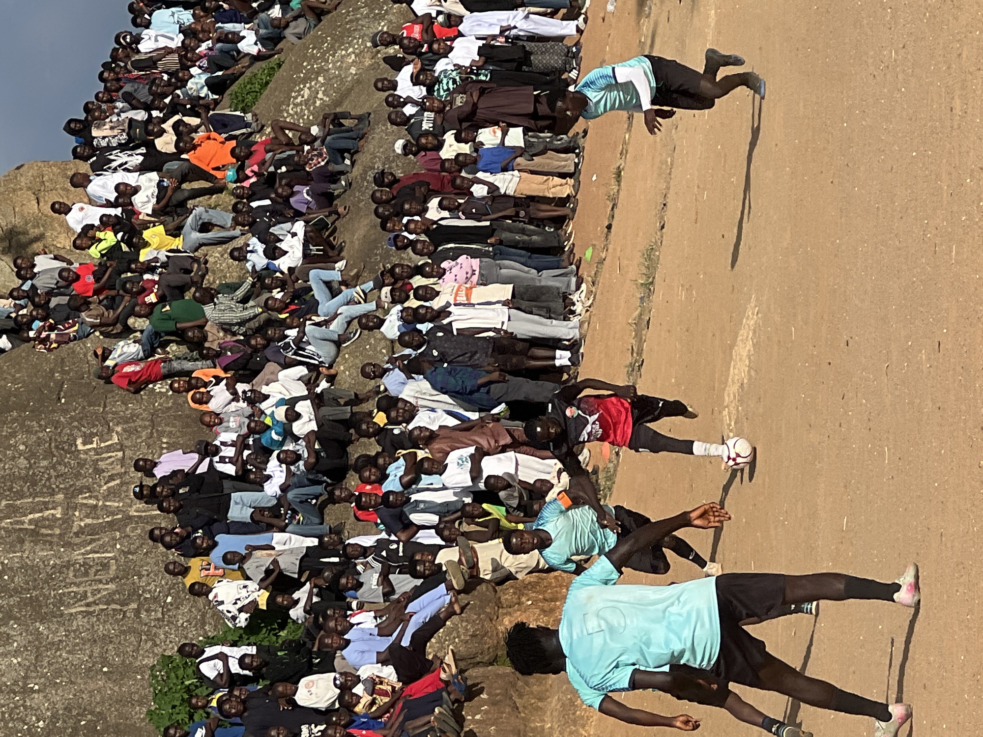 In Jos, Nigeria, a group of young men are playing football on a dusty, outdoor field. The sky is overcast, and a large rocky outcrop with some graffiti is visible in the background, with a crowd of spectators gathered on it. The players are dressed in athletic wear, and the game appears to be in progress, with one player kicking the ball. The scene suggests a lively community event, possibly a local tournament or informal game, drawing attention from a significant number of onlookers. The environment is characterized by dry, reddish-brown earth, typical of the terrain in Jos.