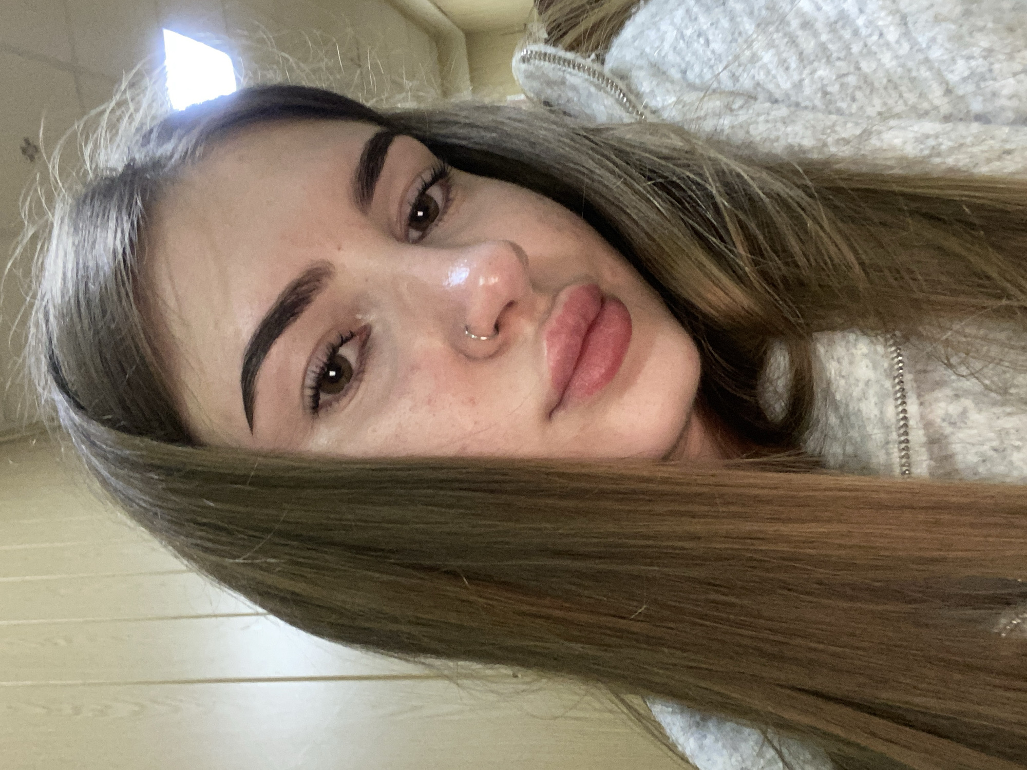 A close-up, rotated portrait captures a young woman looking directly at the viewer. Her long, dark brown hair, with subtle lighter highlights, cascades across the frame. She has well-defined, dark eyebrows, visible eyelashes, and brown eyes. A small, silver-toned ring piercing is visible on her left nostril. Her lips are full, appearing naturally pink and glossy. Her complexion shows some scattered freckles around her nose and cheeks. She is wearing a light-colored, possibly grey or off-white, hooded garment with a visible zipper on the right side of the image.
The image suggests a selfie or a close-up indoor shot, given the proximity to the subject and the visible background elements. The background is softly blurred but reveals light-toned horizontal paneling or a wall surface to the left and bottom. A very bright, possibly overhead, light source is prominent in the upper left corner, creating significant highlights on her forehead and nose, suggesting strong artificial lighting indoors. Her expression is neutral and direct. No text is visible in the image.