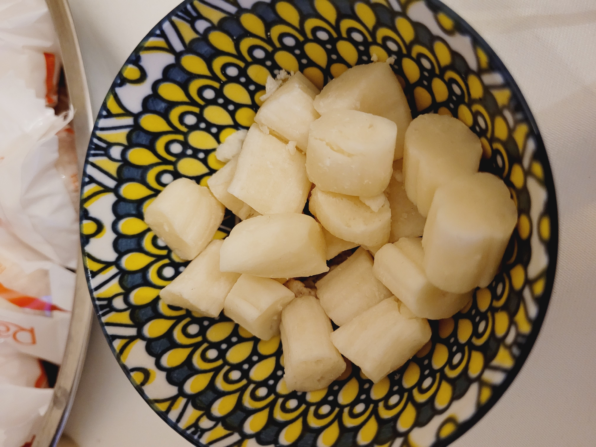 A close-up, overhead shot shows a small, decorative bowl filled with cubed white food. The bowl has a colorful, geometric pattern of yellow, black, and white. The food appears to be some kind of root vegetable, possibly taro or cassava, cut into roughly one-inch cubes. The cubes are slightly irregular in shape and have a smooth, pale surface with some subtle texturing. They are piled loosely in the bowl, with some pieces overlapping. In the blurred background, a silver platter holds several white packets, some with red lettering, suggesting they might contain snacks or condiments. The overall scene is brightly lit, with a white tablecloth providing a clean and simple backdrop. The image does not provide any information about the location in Baku City, Azerbaijan.