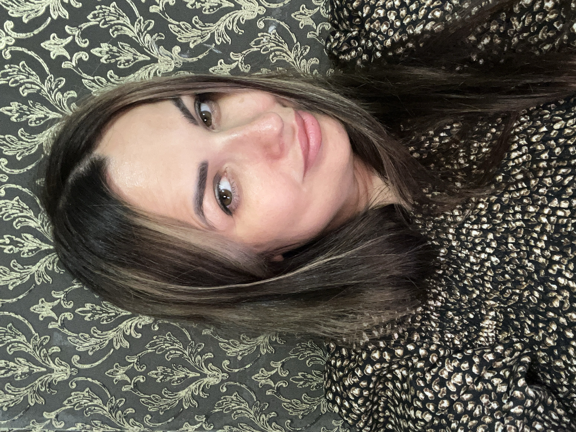 A medium close-up shot shows a woman with long, dark brown hair with blonde highlights, wearing a black and tan animal-print blouse. She is looking slightly to her right with a subtle smile. The background features a dark gray wallpaper with a metallic gold floral damask pattern. The lighting is soft, highlighting the woman's face and the texture of the wallpaper and her clothing.
