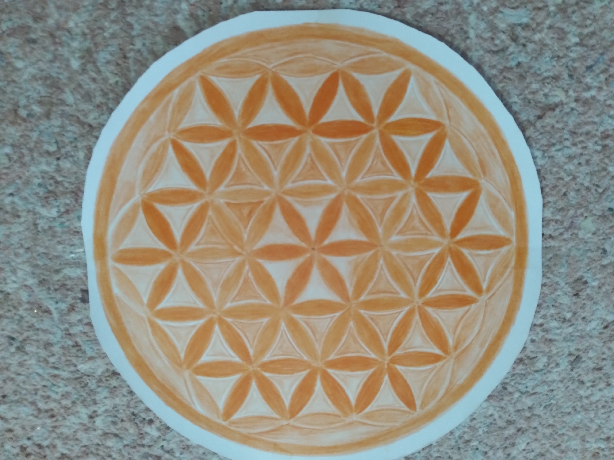 The image shows a circular drawing of the Flower of Life symbol, rendered in shades of orange and white. The drawing is centered on a speckled, sandy-colored surface. There is no visible indication of any specific location in the city of Baki, Uzbekistan, or any other urban setting. The image focuses solely on the artwork and its immediate background.