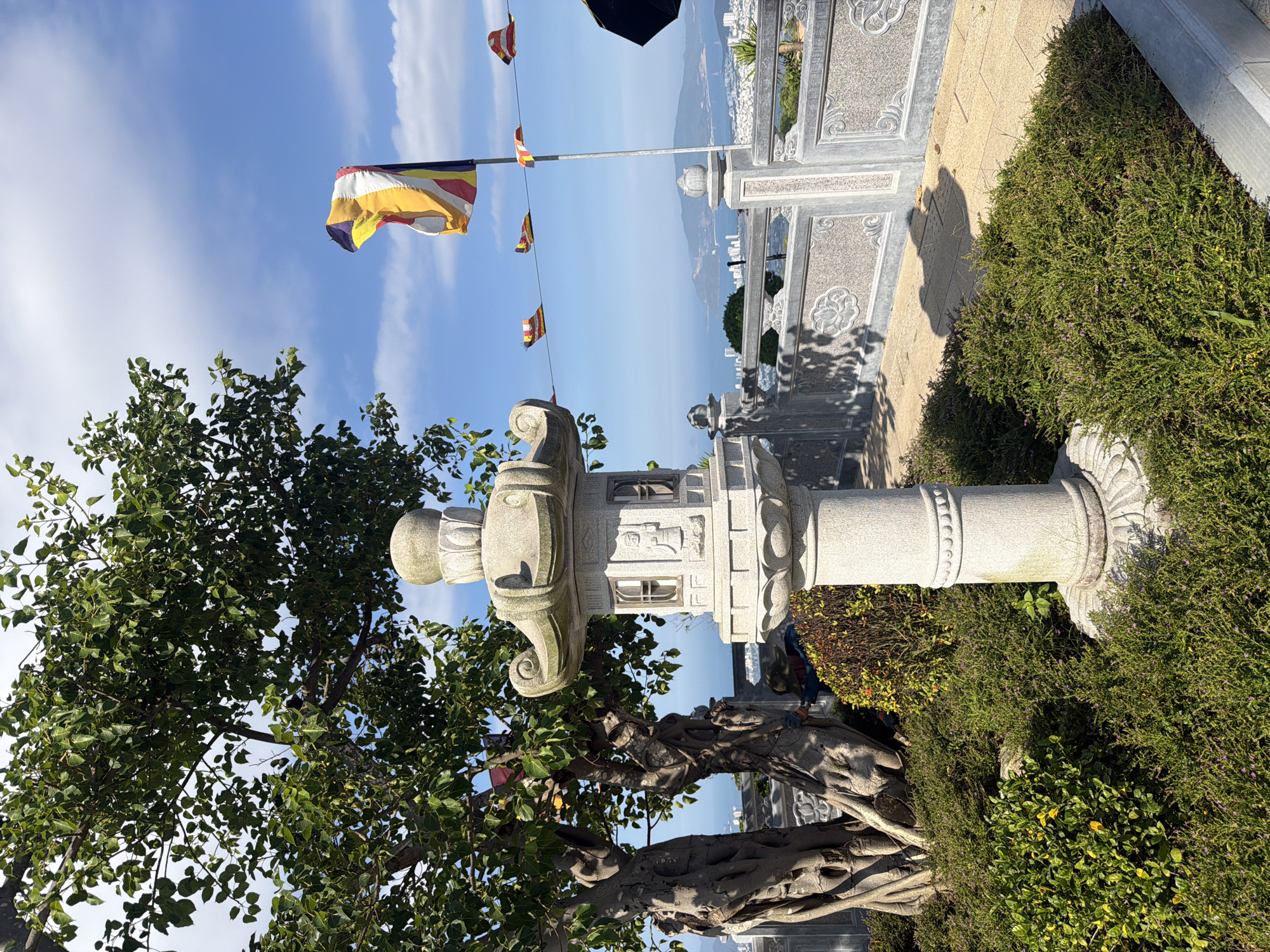 The image depicts a scene in Nam Nha Trang Ward, Vietnam. A stone pillar with ornate carvings stands prominently, with a Bodhi tree beside it. A Buddhist flag is hoisted on a pole in the background. Below the flagpole are more rows of Buddhist flags. A stone railing and a glimpse of the sea and cityscape are visible in the distance. Green foliage surrounds the base of the pillar. The sky is blue with scattered clouds.