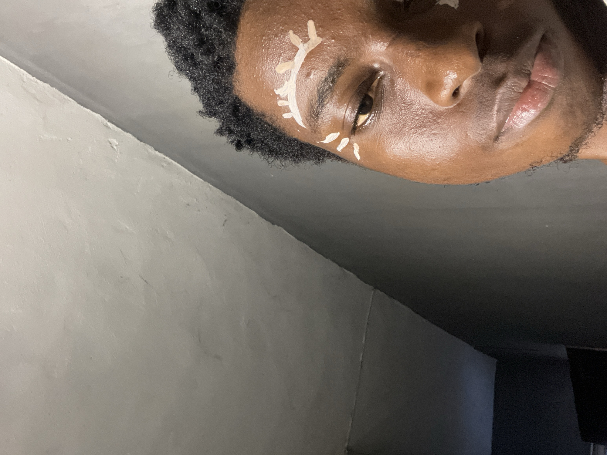 In Nkpor, Nigeria, a close-up, eye-level shot captures the right side of a Black person's face, focusing on their facial makeup. The individual has dark, curly hair and is looking towards the right. White makeup has been applied to their forehead in a stylized design resembling eyelashes. Additionally, white markings are present below their right eye. The background consists of textured, light-colored walls that transition to a darker wall. The lighting is somewhat dim, with a slight sheen on the person's skin, indicating oily or moist conditions.