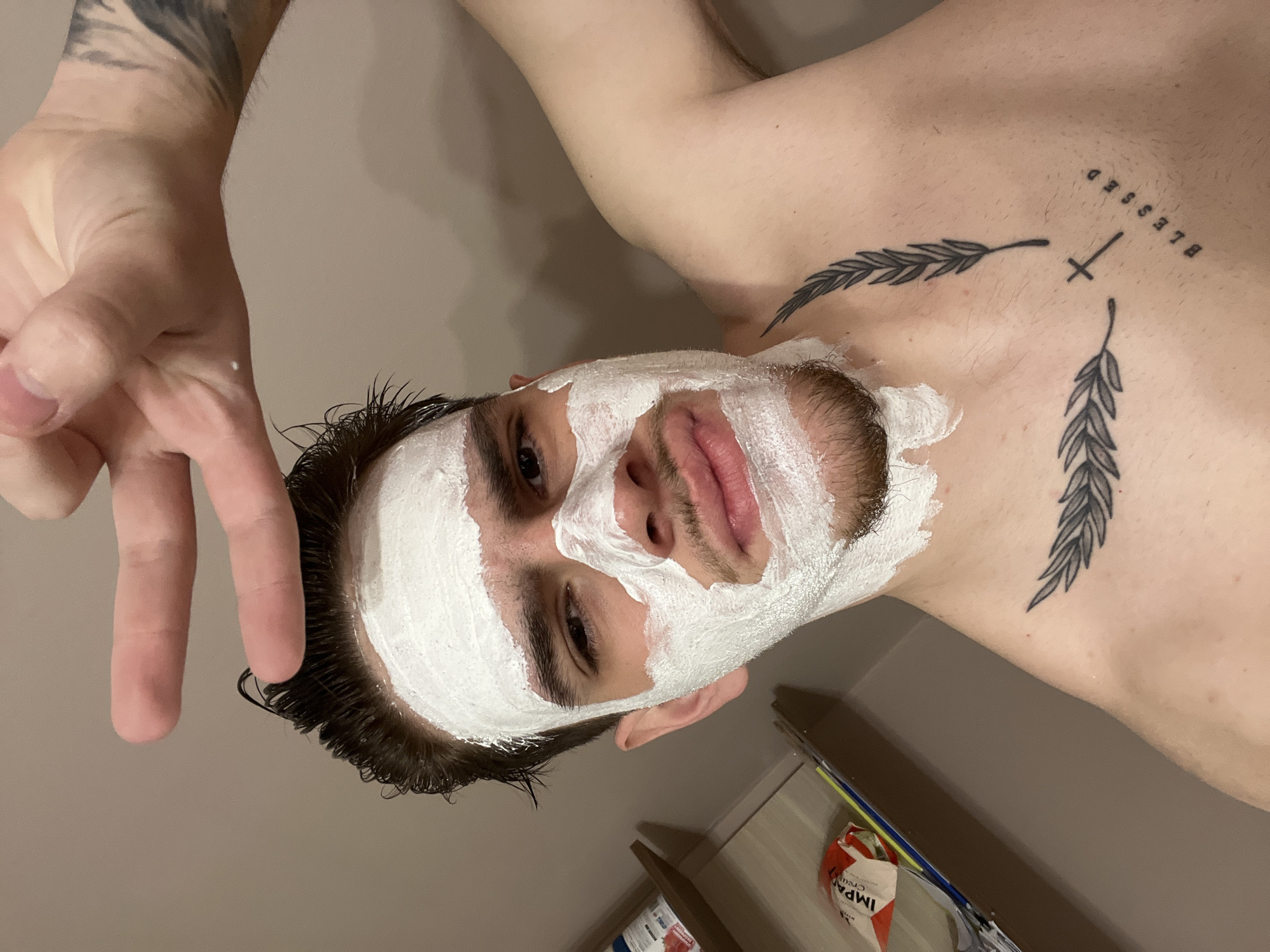 A chest-up, rotated selfie shows a light-skinned man with dark hair and a full white face mask. The mask covers his forehead, cheeks, nose, and the upper portion of his beard. He's making a peace sign with his left hand near his face. Tattoos of feathers and the word "BLESSED" with a cross are visible on his chest and collarbone. The background is a beige wall. The image was not taken in Sofia, Bulgaria.