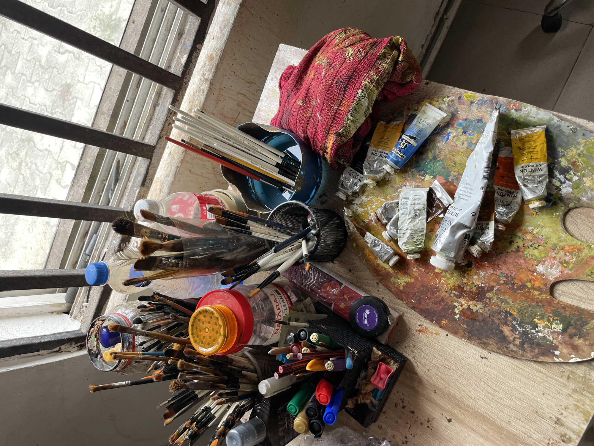 The image depicts an artist's workspace, possibly in Ado, Nigeria, cluttered with art supplies. A wooden table is covered with an assortment of paintbrushes, tubes of oil paint, pens, pencils, and other stationery items. A wooden palette with dried paint is prominently featured, showcasing a vibrant mix of colors. Various containers hold brushes, including a glass jar filled with water and brushes, and a metal mesh cup. Among the items are bottles of water and a red fabric. The scene is set near a window with metal bars, suggesting an indoor environment. The overall impression is one of a well-used and creative space.