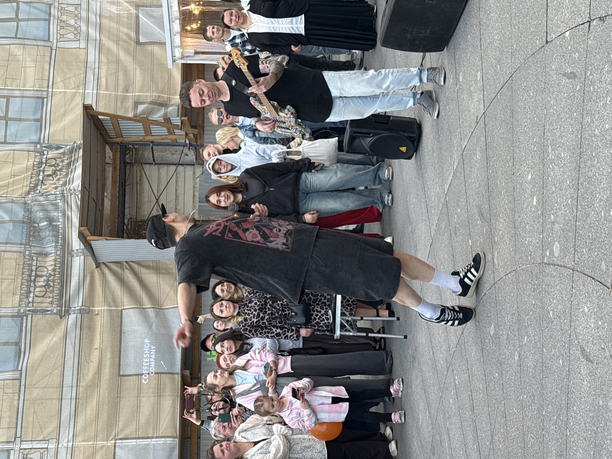 A group of people are gathered outdoors, with a performer on a bass guitar in the foreground, playing for the crowd. A second performer, wearing a cap and dark clothing, stands to the left, pointing towards the crowd while holding a microphone. Behind them, a backdrop depicts a building facade, possibly a mural or a projection, with the words "COFFEESHOP COMPANY" visible. The gathering appears to be taking place on a paved public space, likely a street or square in Saint Petersburg, Russia. The audience consists of individuals of various ages, some holding phones to record. The atmosphere seems to be that of a casual outdoor performance or event.