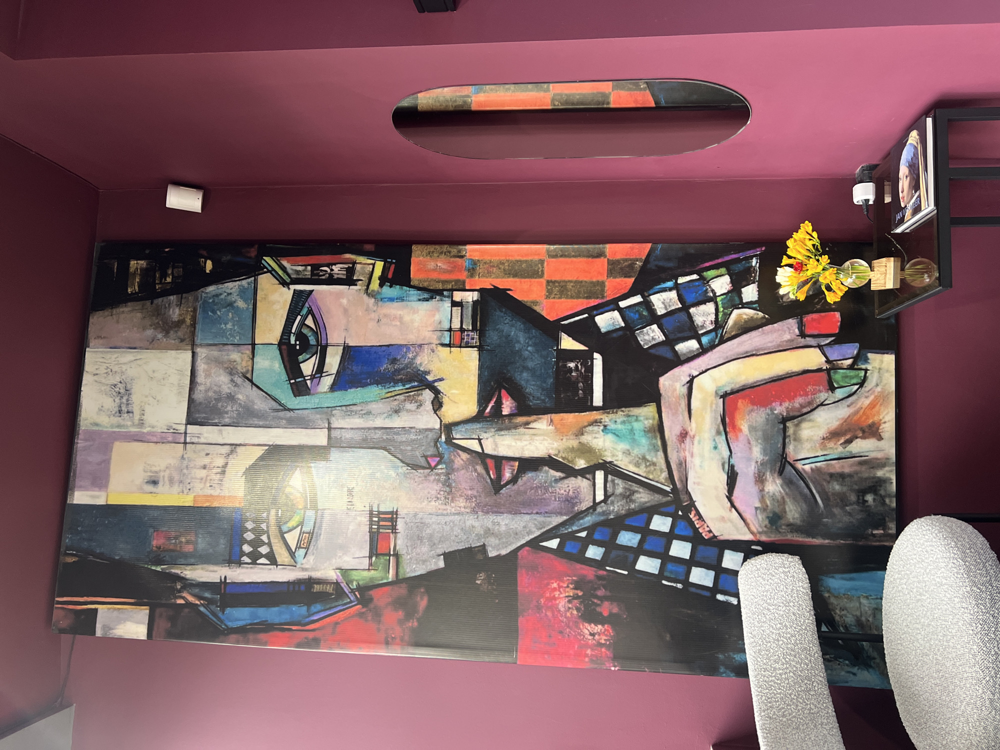 The image shows an interior space in Yerevan, Armenia, dominated by a large, abstract painting on the wall. The painting features a collage of geometric shapes and fragmented figures in a Cubist-inspired style, with a checkerboard pattern incorporated in the background.
The walls and ceiling are painted in a dark, purplish-red color. A rectangular, recessed light fixture is visible in the ceiling above the painting.
To the right of the painting, a black metal shelf unit holds a framed print of Jan Vermeer's "Girl with a Pearl Earring", a lightbulb and a small bouquet of yellow and red flowers in a vase.
Two tall, white, textured chairs are partially visible in the foreground on the right side of the image, suggesting a seating area. A small white device is attached to the wall above the left side of the painting.