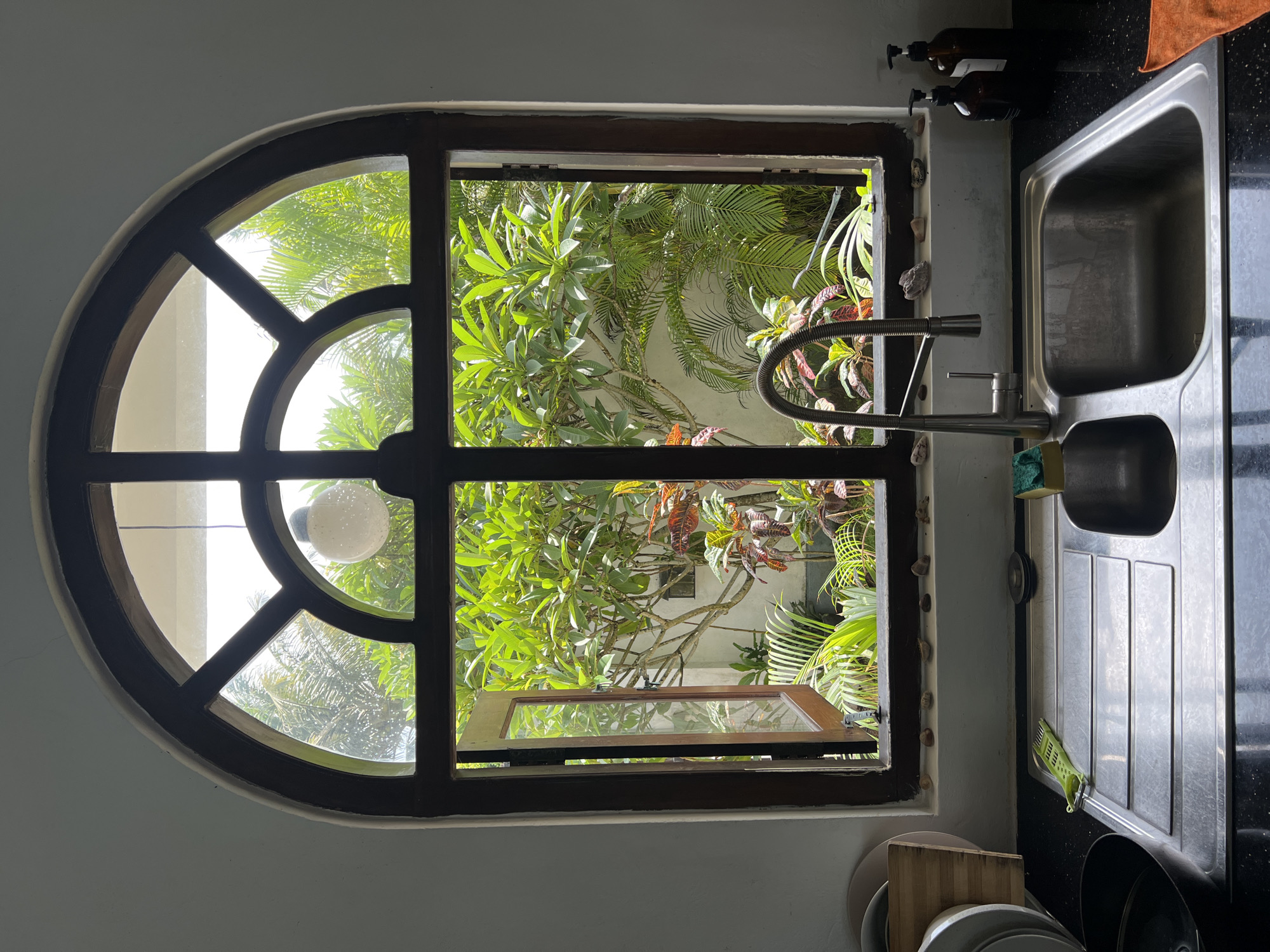 A view from a kitchen sink in Goviyapana, Sri Lanka, reveals a window with a rounded top and a rectangular lower section, partially open. The window frames a view of dense tropical foliage. The kitchen sink is stainless steel, with a modern-style faucet. Dishes and cutting boards are visible below. Two bottles with pump dispensers are visible next to the sink on the left.
