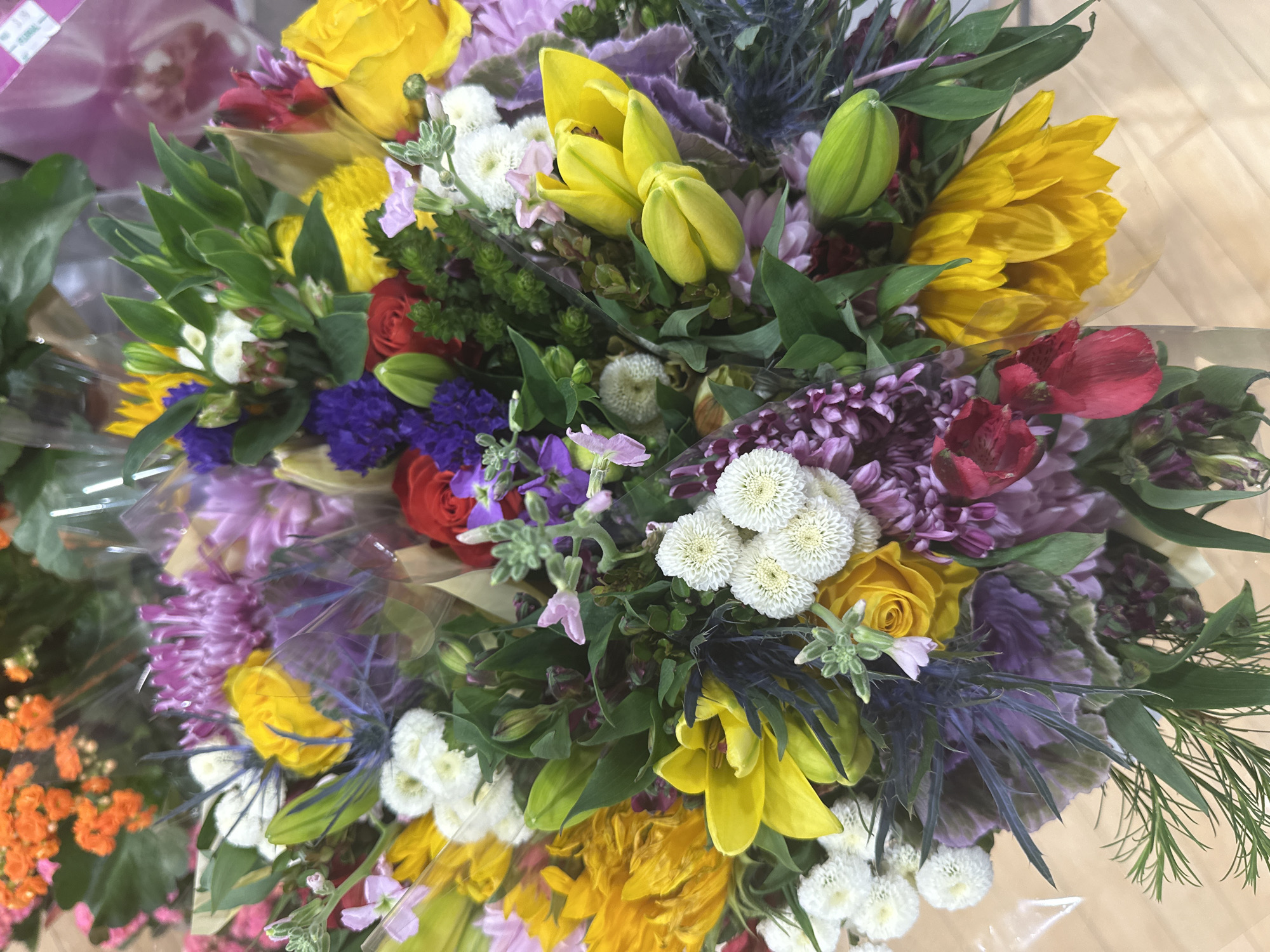 The image shows a close-up, overhead view of a colorful bouquet of flowers. The bouquet is arranged with a variety of flowers in shades of yellow, purple, white, red, and orange. Some of the prominent flowers include sunflowers, lilies, roses, and mums. Green foliage and some purple thistles add texture and depth to the arrangement. The flowers appear to be fresh and are wrapped in clear plastic, suggesting they are intended for sale or as a gift. The background is blurred but appears to be a light-colored surface, possibly a table or countertop. The scene is indoors, and no people are visible. The specific location within Camp Hill, United States of America, is not discernible from the image itself.