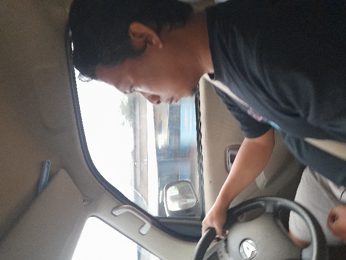 A male individual is depicted driving a vehicle in Temuwulan, Indonesia. He is positioned in the driver's seat, viewed from the chest up, wearing a black short-sleeved t-shirt with a light design on the left chest and light-colored pants. A light-colored seatbelt crosses his chest. His right hand grips the steering wheel, which features a silver Daihatsu emblem. His head is turned slightly to the right, with his gaze directed forward. The vehicle's interior is light-colored, showing a sun visor in the upper left and an A-pillar grab handle. Through the driver's side window, a blurred exterior environment indicates the vehicle is in motion, revealing an elevated concrete structure or road, green foliage, and a blue object in the distance under a bright sky.