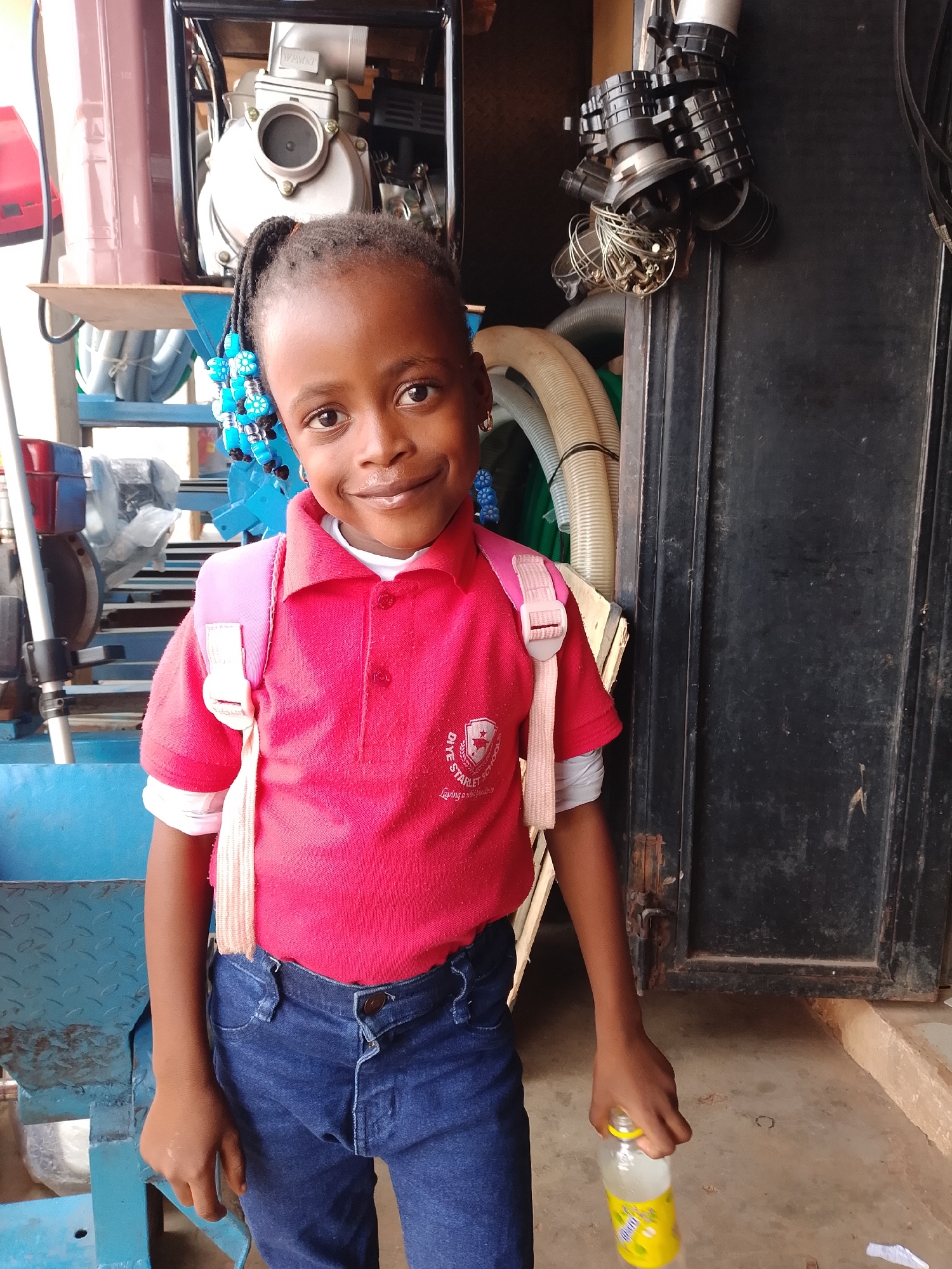 A young girl stands smiling, facing the camera, in what appears to be an indoor or covered industrial-like setting. She has dark skin and braided hair adorned with blue beads, and is wearing earrings. She is dressed in a bright red short-sleeved polo shirt, with a white long-sleeved shirt underneath, and blue denim jeans. A pink backpack with white straps is visible on her back. In her right hand, she holds a clear plastic bottle with a yellow cap and a yellow and white label, which appears to have the text "VIT" and possibly "Nimbo" or similar branding.
The background is cluttered with various objects. To the left and behind her, there is a light-colored machine, possibly a water pump or engine, resting on a blue metal stand. A large, brown cylindrical container is also visible. To her right, a dark, possibly metal, structure or door frame is present, from which several industrial components, including dark, ribbed connectors and tangled wires, hang. Further back, bundles of white or beige corrugated hoses and pipes are stacked.
On the left chest of her red polo shirt, there is a visible white logo with a star and a shield, accompanied by the text "GIVE STARLET SCHOOL". Below this, smaller text is partially obscured but appears to say "Laying a solid foundation". The floor is a light-colored concrete. The lighting suggests it is daytime. The girl's cheerful expression is a notable emotional detail.