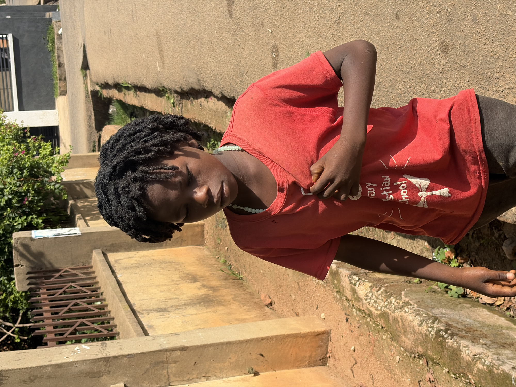 A young person with dark skin and dreadlocks, wearing a red t-shirt and a light green beaded necklace, stands near concrete steps. The t-shirt has white writing and a graphic on the front. The background shows vegetation and urban architecture. The scene appears to be in an outdoor area. Given the prompt, this may be the city of Jos, Nigeria.