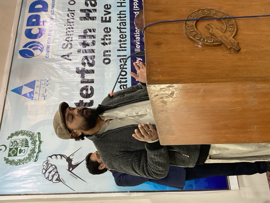 An indoor scene at the Quetta Press Club in Quetta City Tehsil, Pakistan, depicts a male speaker addressing an audience. The primary subject, a man with a beard, a patterned flat cap, a beige collared shirt, and a grey zippered cardigan, stands behind a wooden podium. His hands are raised and open, palms forward, indicating a communicative gesture. A black microphone with a red tip is attached to the podium's front. The podium, exhibiting signs of wear, features a carved "QUETTA PRESS CLUB" logo on its lower right side. Behind the speaker, a large white banner serves as a backdrop, displaying the Government of Pakistan emblem, an illustration of two clasped hands, and logos for "PPAF" (Pakistan Poverty Alleviation Fund) and "CPD" (Centre for Peace & Development). Visible text on the banner indicates "A Seminar on Interfaith Ha..." and "...on the Eve of International Interfaith Ha...", with a credit to "Alleviation Fund (PPAF)". A second male, wearing a dark suit jacket, is partially visible behind the main speaker to the left.