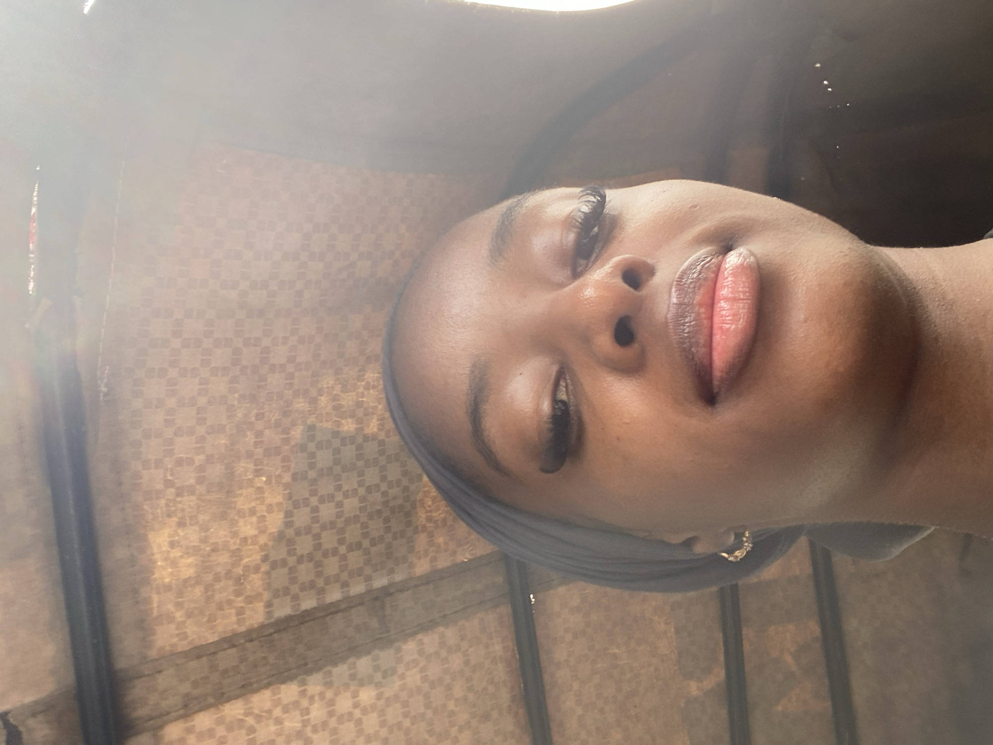 This media file is a portrait of a young Black woman, likely taken indoors or in a covered outdoor space, given the patterned material visible in the background. The image is oriented vertically, with the woman's face dominating the frame. She is wearing a blue headscarf and a hoop earring in her left ear. Her makeup includes pronounced eyelashes and lip gloss.
The lighting suggests it could be daytime, with a bright light source creating a strong highlight on her forehead and the left side of her face. The background material has a repetitive, checkered or woven pattern in earthy tones, possibly a fabric or a textured wall covering. The overall mood of the image is relaxed and intimate, focusing on the subject's features. There is no visible text in the image. The location context provided is Jos, Nigeria.