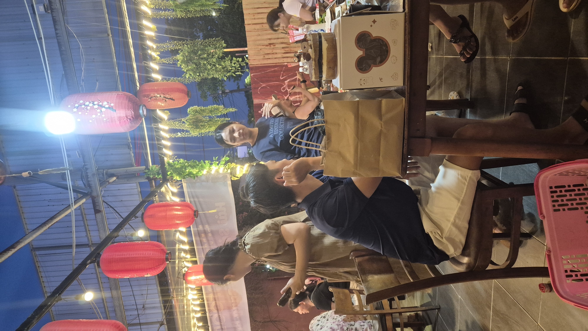 In Đà Nẵng, Vietnam, a group of people are gathered at an outdoor restaurant during twilight. Red lanterns hang from the ceiling, illuminating the scene. A man in a dark shirt and white shorts sits at a table, looking to his right. A woman in a brown dress stands beside him, holding a phone. Another woman in a navy blue t-shirt stands behind the man, looking towards the camera. A cake sits on the table, decorated with frosting and chocolate. A brown paper bag is also on the table. The floor is tiled, and plants hang from the ceiling. The overall atmosphere appears to be a casual gathering, possibly a birthday celebration.