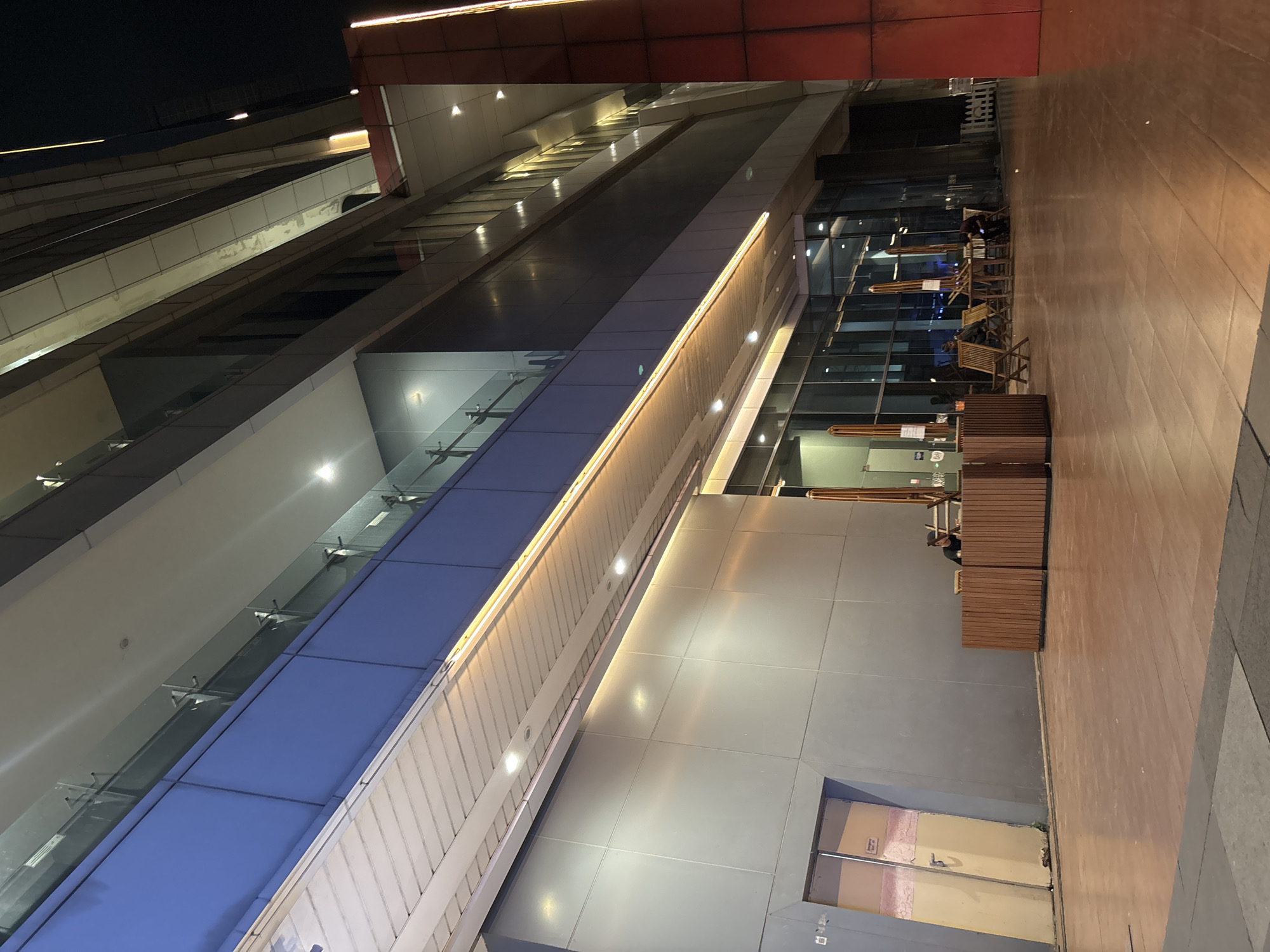 The image depicts the exterior of a modern building at night, likely a commercial or office complex, located in North Jakarta, Indonesia. The building features a sleek, multi-story facade with extensive glass windows and metallic paneling. A covered walkway with a slatted ceiling and strip lighting runs along the ground level, illuminated by overhead lights and a warm-toned band of illumination along the edge.
In the foreground, a polished wooden walkway or patio extends from the building. To the left, a pale yellow door with visible scuff marks and a metal frame is ajar, revealing a dimly lit interior. To the right of the door, a person is seated behind a wooden counter-like structure. Further to the right, several outdoor seating arrangements are visible, including wooden chairs and tables, some with umbrellas. A few individuals are seated at these tables, one appearing to be looking at a mobile device. The overall environment suggests a commercial area that is either closing or experiencing a slow evening. The lighting is artificial, creating a sense of depth and highlighting the architectural features of the building.