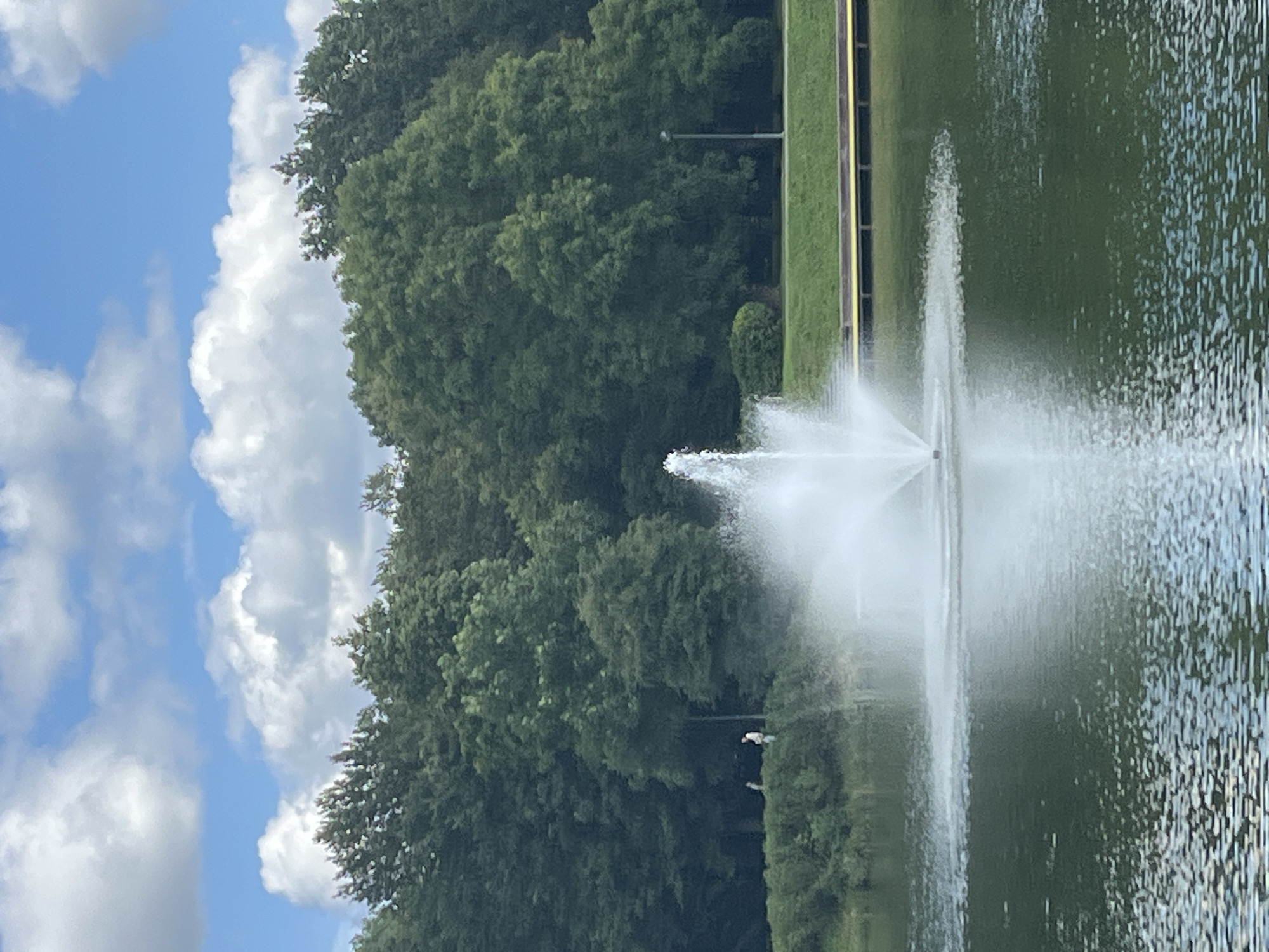 The image shows a fountain spraying water in a lake, with a dense line of trees in the background and a bright blue sky with white clouds above. There are two people standing on the grassy bank near the trees, observing the fountain. The lake is calm, with ripples on its surface. The scene appears to be a park or recreational area. There is no information in the image to suggest it is located in Kohtla-Järve linn, Estonia.