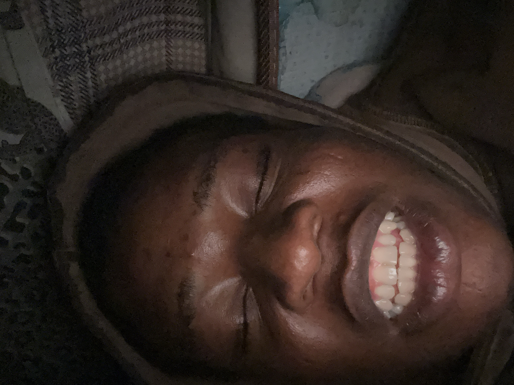 A close-up, low-angle shot depicts a person with dark skin, lying down with their eyes closed and a wide smile revealing their teeth. The person appears to be wearing a hooded garment. The surrounding environment includes patterned fabrics that suggest bedding or clothing. The location is not specified or discernible from the image.