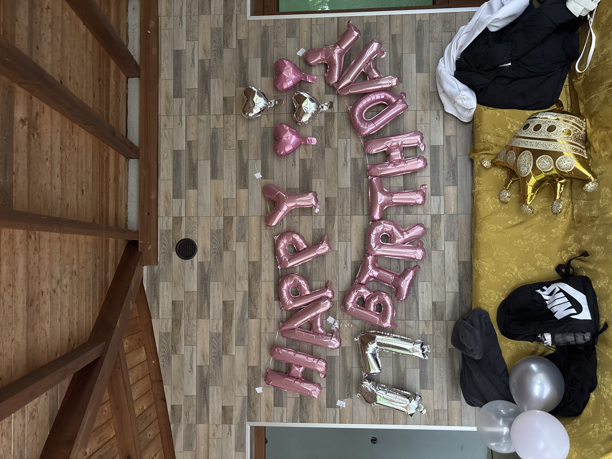 The scene appears to be an indoor space, possibly a room within a house or apartment. There is wood flooring. A "Happy Birthday" balloon display, consisting of pink and silver letter and heart-shaped balloons, is spread out on the floor. Adjacent to the balloons is a couch or bed covered in yellow fabric. On the couch are clothing items, including a white and black jacket, a gold crown-shaped balloon, a black Nike backpack, gray sweatshirts, and silver balloons. The presence of the celebratory balloons and other items suggests a birthday celebration. The angle of the picture indicates that it was taken from above. Based on the inclusion of Zlatograd, Bulgaria, this scene is likely located there.