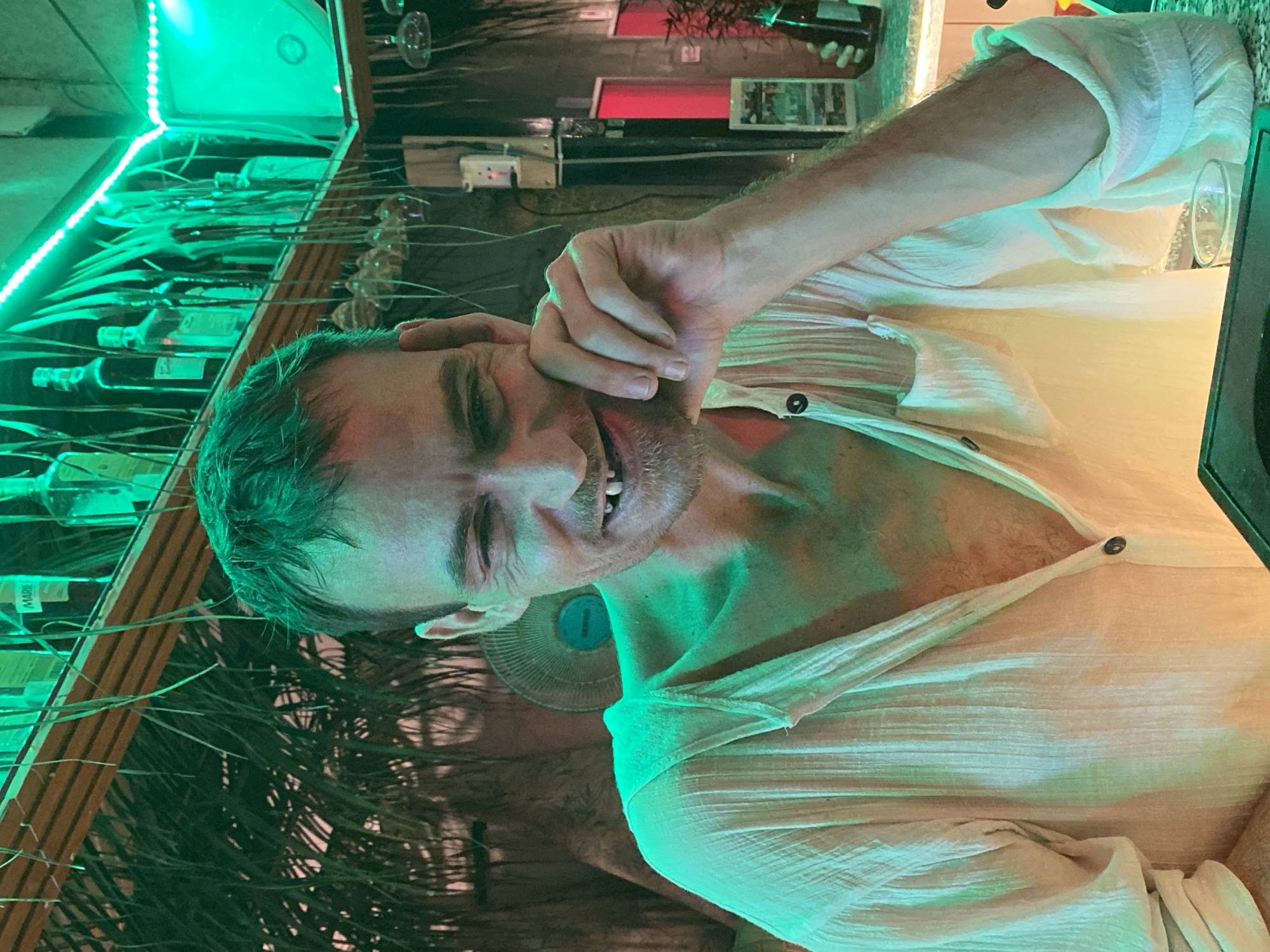 An adult male with short dark hair, stubble, and a visible gap in his upper front teeth smiles and winks, resting his left hand on his cheek. He is wearing a light-colored, textured, long-sleeved shirt with buttons, unbuttoned to reveal chest hair. The scene is illuminated by prominent green neon lighting, casting a green hue across the man and the environment.
The location is a bar or restaurant in Mũi Né, Vietnam. In the background, behind the man, a bar setup is visible. Above his head, a shelf displays various liquor bottles, some dark and some clear, with a green LED strip light running along its edge. Below the shelf, numerous dark, thin, leafy fronds or decorative foliage hang down. To the man's right (viewer's left), more foliage is present. Behind his right shoulder, a light-colored circular fan is mounted on the wall. To his left (viewer's right), several stemmed glasses hang inverted from a rack. Further to the right, a power outlet is visible on a dark post. Red rectangular elements are seen in the far background. On the counter to the right, a small plant in a dark pot and a display stand with images are present. A clear glass containing liquid sits on a dark counter surface in the foreground. The counter surface beneath the hanging glasses appears to have a light, patterned texture.