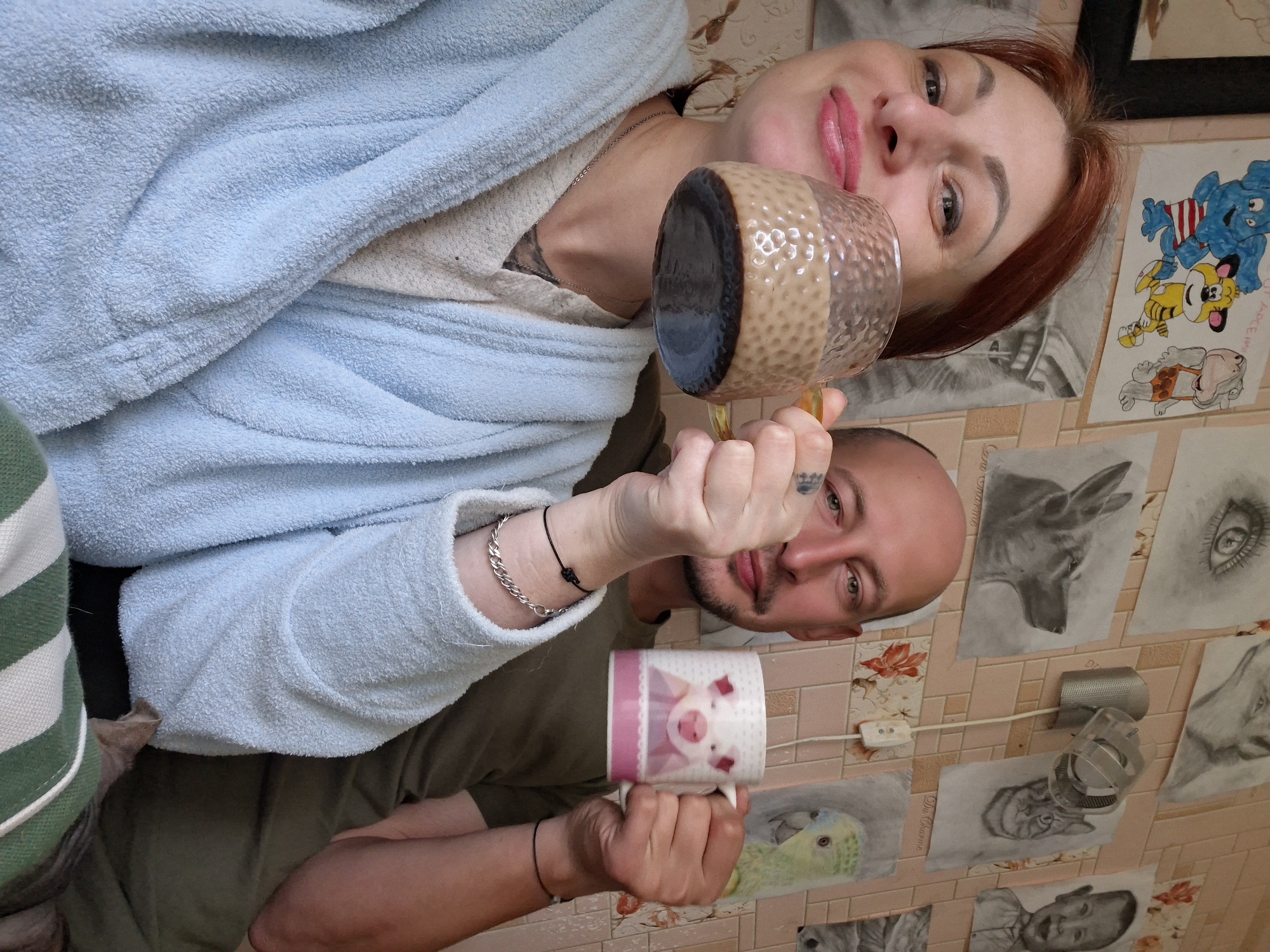 A medium shot captures a couple holding mugs, positioned centrally against a wall adorned with various framed drawings. The woman, on the left, is in the foreground and slightly out of focus. She has red hair, and her head is tilted to her left as she smiles, holding a clear glass mug filled with a brown beverage. She wears a light blue terrycloth robe over a textured cream-colored garment and a silver chain necklace. On the right, a bald man with a mustache looks towards the camera. He wears a dark olive green t-shirt and holds a white mug decorated with a pink pig. His left arm, bent at the elbow, is visible. Both subjects have tattoos on their hands and arms. The background wall features framed pencil sketches of animals and a human eye, along with some decorative floral wallpaper. A small metallic fixture with a visible wire and plug is mounted on the wall between the drawings. The overall setting appears to be an indoor, domestic environment, likely a room in a house or apartment in the city of Myadzyel, Belarus.