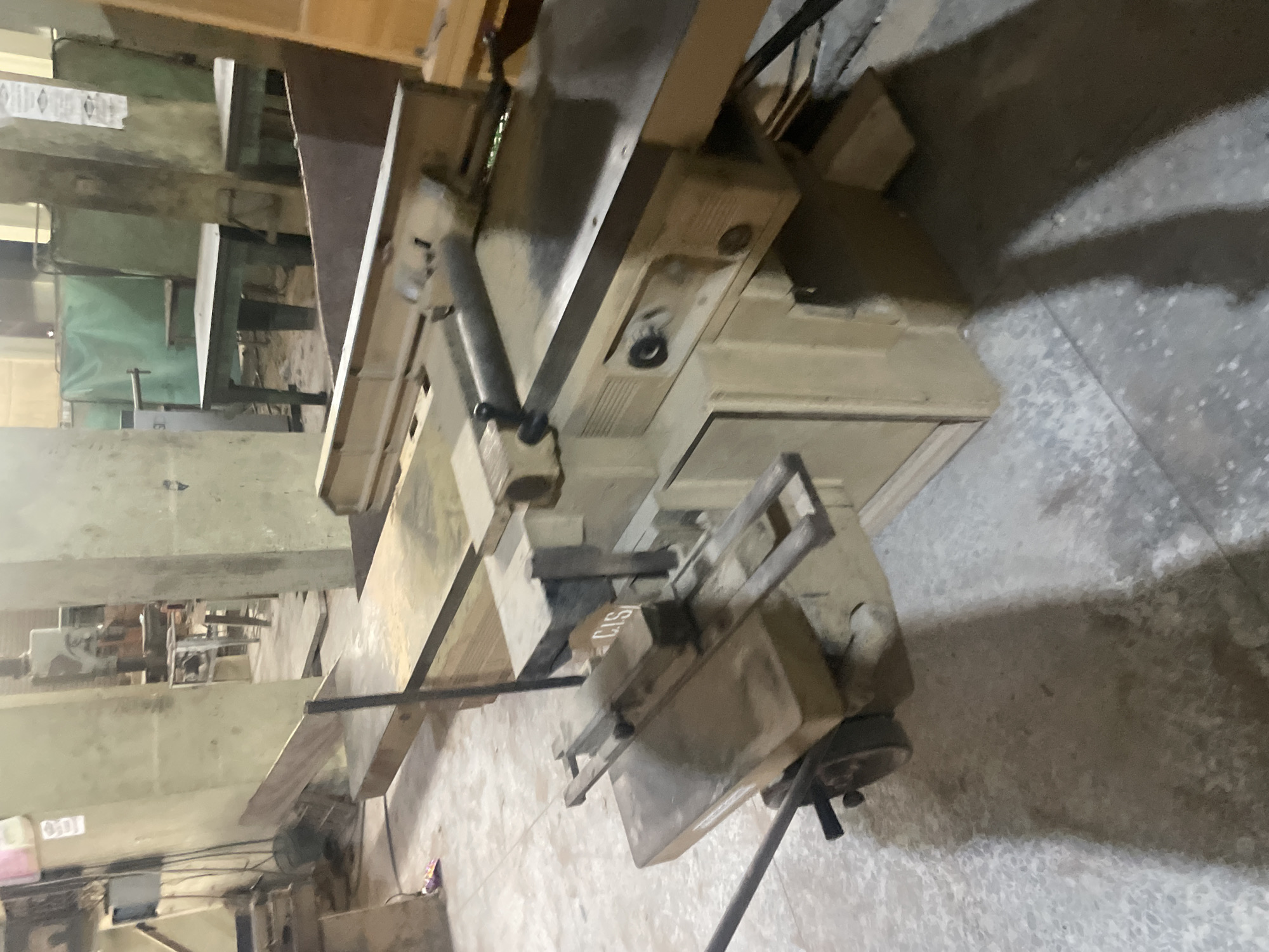 This image depicts a woodworking machine, likely a jointer or planer, situated in what appears to be a workshop or industrial setting in Ihiagwa, Nigeria. The machine is predominantly constructed of metal and wood, with a beige and metallic finish. Key components visible include a long, flat bed, a blade housing with a guard, and various knobs and levers for operation. The machine is positioned on a concrete floor, and shadows cast by the overhead lighting suggest an interior space. In the background, other industrial equipment and materials are visible, including what seem to be workbenches, partitions made of green fabric, and stacks of wood. The overall environment appears somewhat cluttered, consistent with an active workshop. No individuals are present in the immediate vicinity of the woodworking machine, and no specific actions are being performed. The lighting is functional, illuminating the equipment for work.