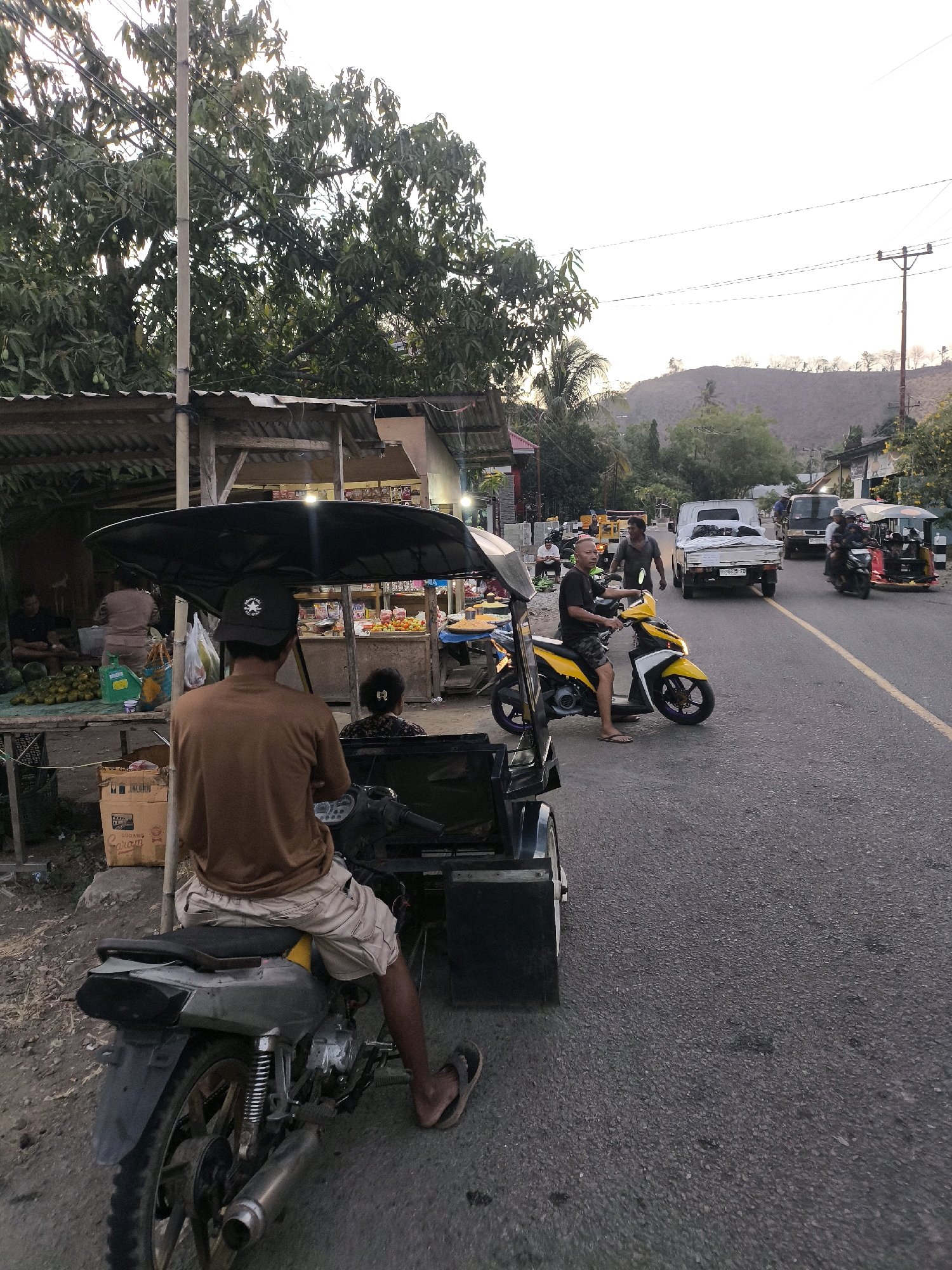 The image depicts a street scene in an Indonesian city at dusk. In the foreground, a person wearing a brown t-shirt and khaki shorts is seated on a motorcycle with a sidecar. They are facing away from the camera. To the left, a market stall is set up with various fruits and goods displayed. Several people are visible around the market, including one person sitting on a stool and another attending to the stall. On the street, other vehicles are present, including a yellow and white scooter with a rider, a white pickup truck, and a yellow auto-rickshaw. The road is paved and has a yellow line marking the center. In the background, there are trees, buildings, and a hillside. The sky is overcast, suggesting the end of the day.