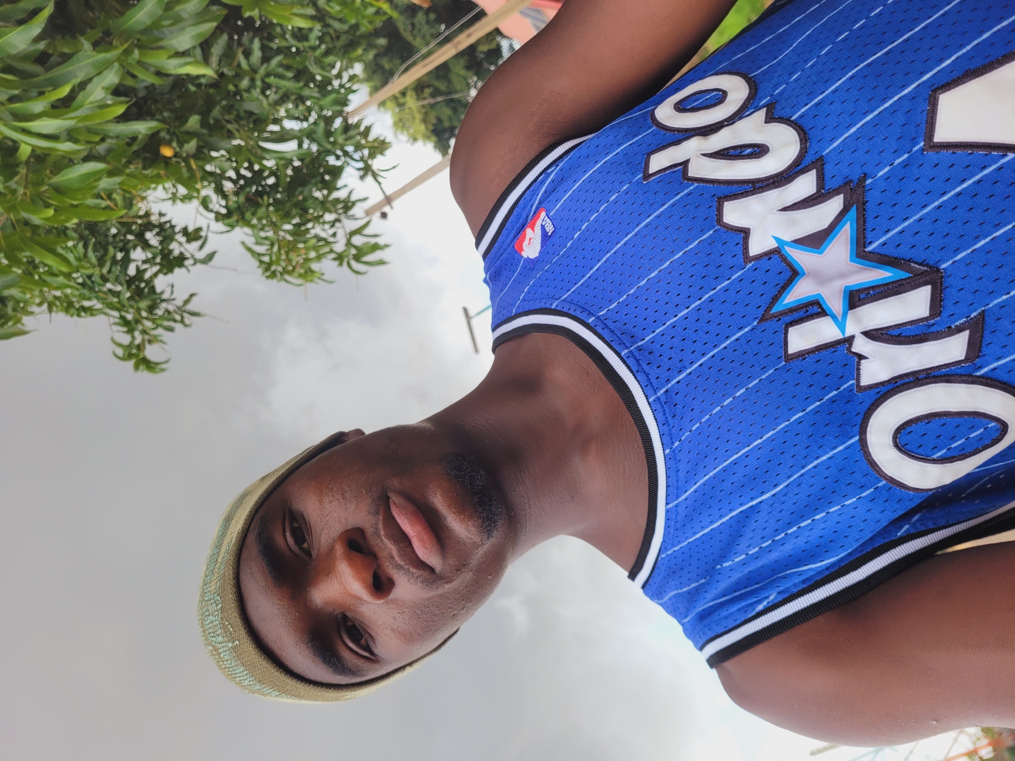 A young Black man is pictured in a low-angle, outdoor selfie. He wears a green beanie and a blue basketball jersey with "Orlando" and a star emblem emblazoned on the front. The jersey appears to be a replica of the Orlando Magic's NBA team. The background consists of a cloudy sky and lush green foliage, suggesting a tropical or subtropical environment. The man is looking towards the camera with a neutral expression. The context of Du, Nigeria is absent from the visual information in the image.