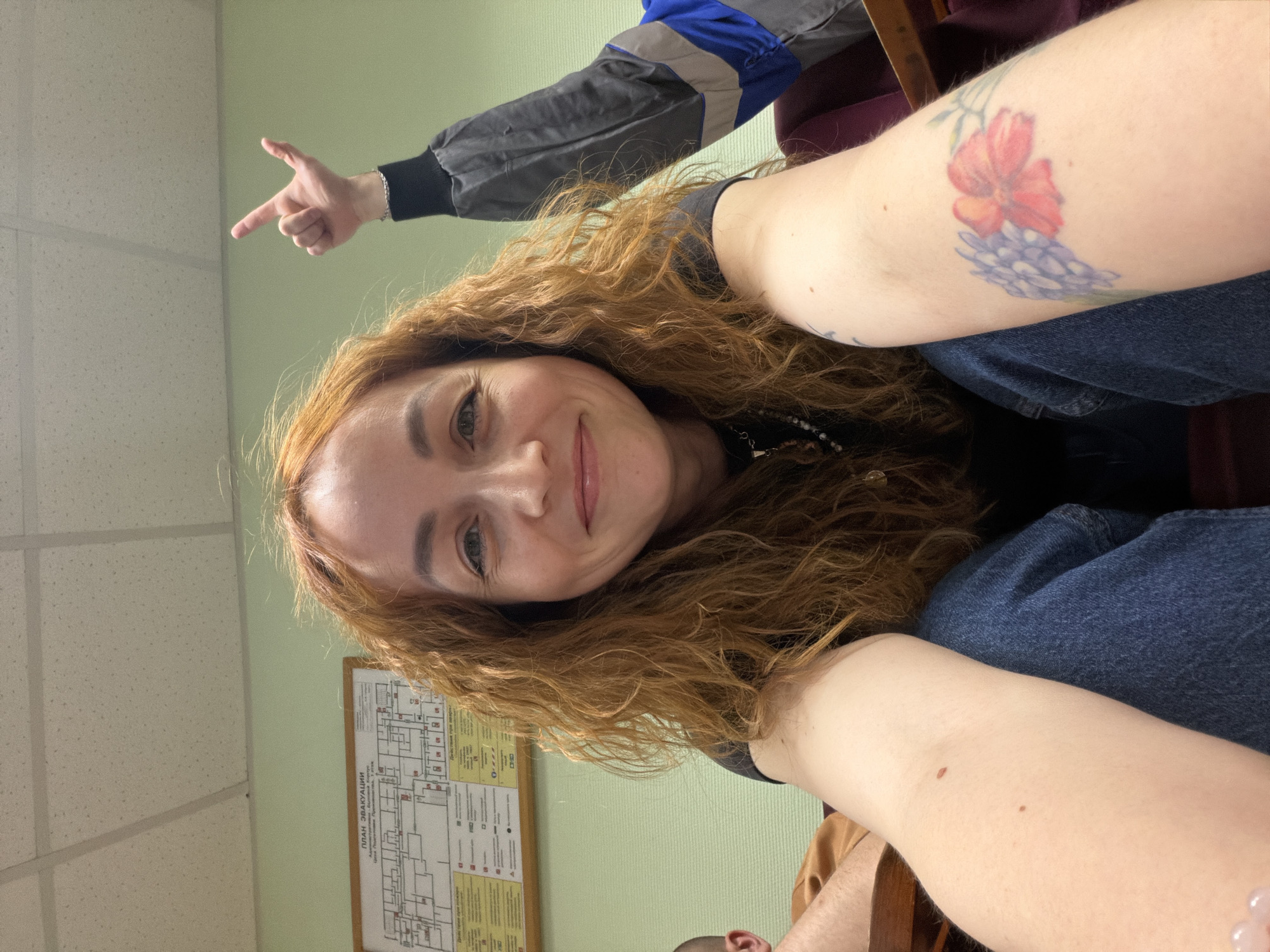 A woman with long, curly, reddish-brown hair is smiling and looking directly at the camera, seemingly taking a selfie in an indoor setting. She is wearing a dark top and blue jeans. On her visible left arm, there are two distinct tattoos: a vibrant red flower and a cluster of purple flowers, resembling lavender or hyacinths.
The background features light green walls and a white acoustic tile ceiling. To the left, an arm wearing a grey sleeve with a blue and reflective stripe is visible, making a pointing gesture. A portion of another person, clad in brown, is also subtly visible at the bottom of the frame. On the wall behind the woman, a framed diagram or chart is mounted. The most prominent text visible on this chart reads "ПЛАН ЭВАКУАЦИИ," which translates to "EVACUATION PLAN" in Russian.
The woman's expression is friendly and pleasant. The presence of the Russian text on the evacuation plan aligns with the provided context of Chaikovsky, Russia, suggesting the image was taken there. The lighting appears to be artificial, typical for an indoor environment.