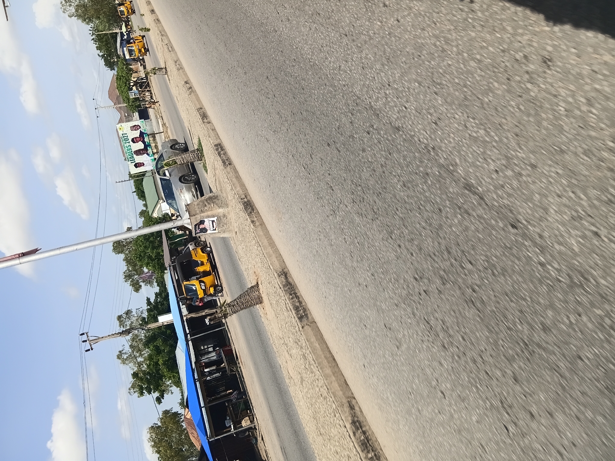 The image shows a street scene in Jalingo, Nigeria. A silver sedan is parked on the right side of the road, facing forward. To the left of the sedan, there are several yellow auto-rickshaws, also known as 'Keke NAPEPs'. In the background, a large billboard displays images of three men and the text "LEADERS 2027". A blue-roofed structure, possibly a market stall or a shop, is visible on the far left. The road is paved and appears to be a busy thoroughfare, with some vegetation and utility poles lining the street. The sky is blue with some scattered clouds.