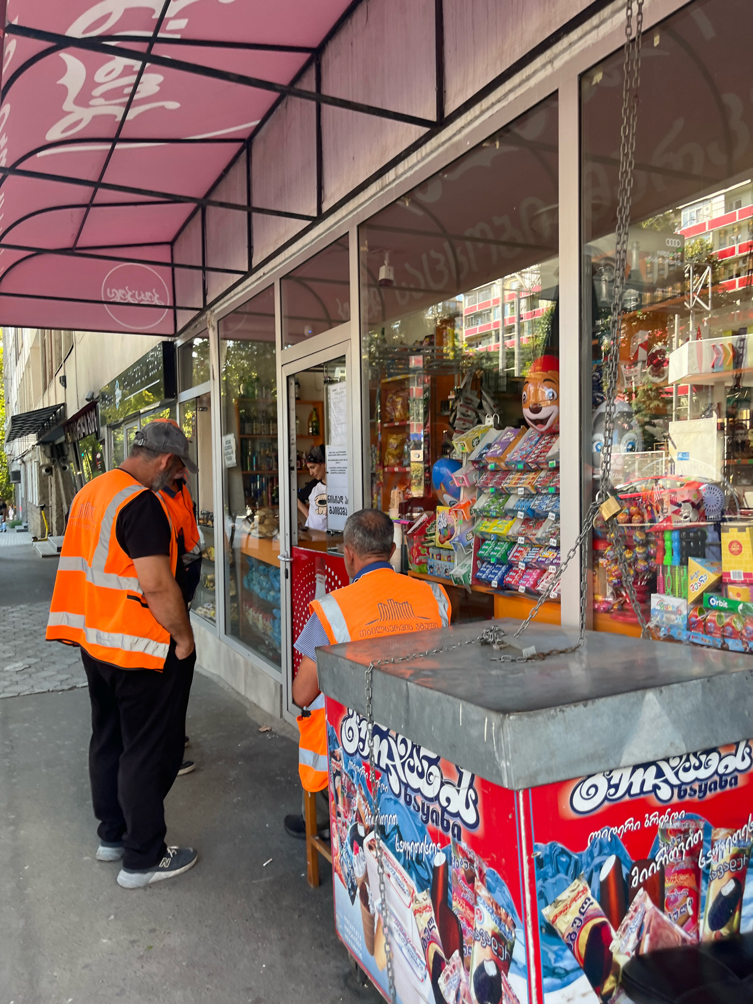 In Tbilisi, Georgia, on a sunny day, three men wearing orange safety vests stand on a sidewalk in front of a small storefront that appears to be a convenience store or a kiosk selling snacks and ice cream. Two of the men are facing the store, while one is facing away from the camera. The store has large glass windows displaying various products, including packaged snacks, drinks, and ice cream. There is a pink awning above the entrance, and a metal chain hangs down in front of the window, secured with a padlock. A red and silver ice cream cooler with images of ice cream cones is positioned on the sidewalk in front of the store. In the background, residential buildings and trees are visible. The overall scene suggests a commercial area within the city.
