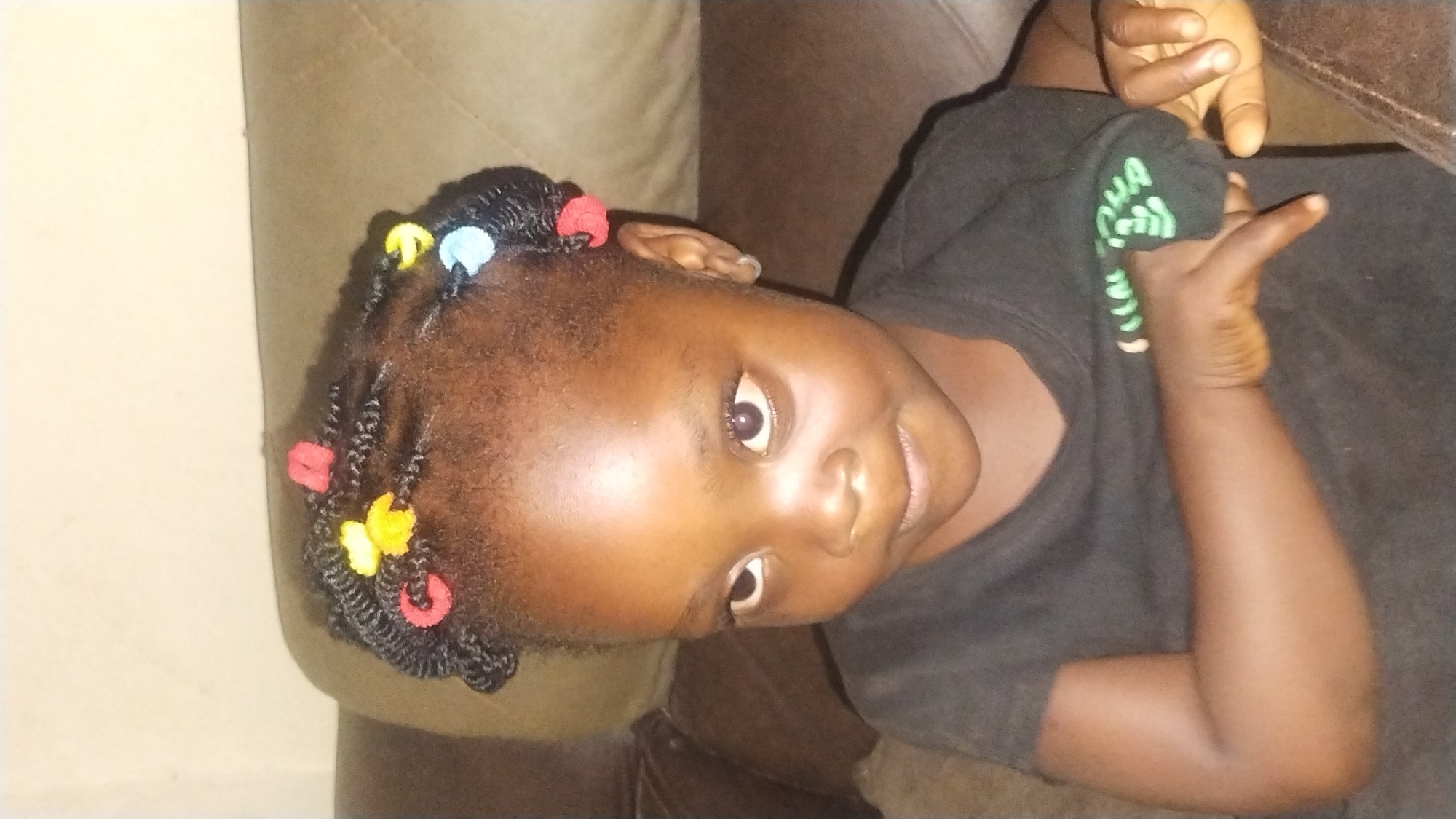 This image captures a young child, appearing female, with dark skin, situated indoors in what is contextualized as the city of Jos, Nigeria. The child is viewed from the chest up, looking slightly upwards and to the viewer's left. The child's hair is dark, styled into numerous small, tightly coiled twists adorned with multiple colorful hair accessories in red, yellow, and light blue. A small, silver-colored hoop earring is visible in the child's right earlobe. The child wears a dark-colored t-shirt, with discernible light green text, including the letters "OHA," on the left sleeve. The child's left arm is bent across the torso, with the right hand positioned nearby. The background features a light-colored wall above and a brown, upholstered surface, consistent with a sofa or couch, behind and to the sides of the child. The scene is illuminated by ambient indoor light.