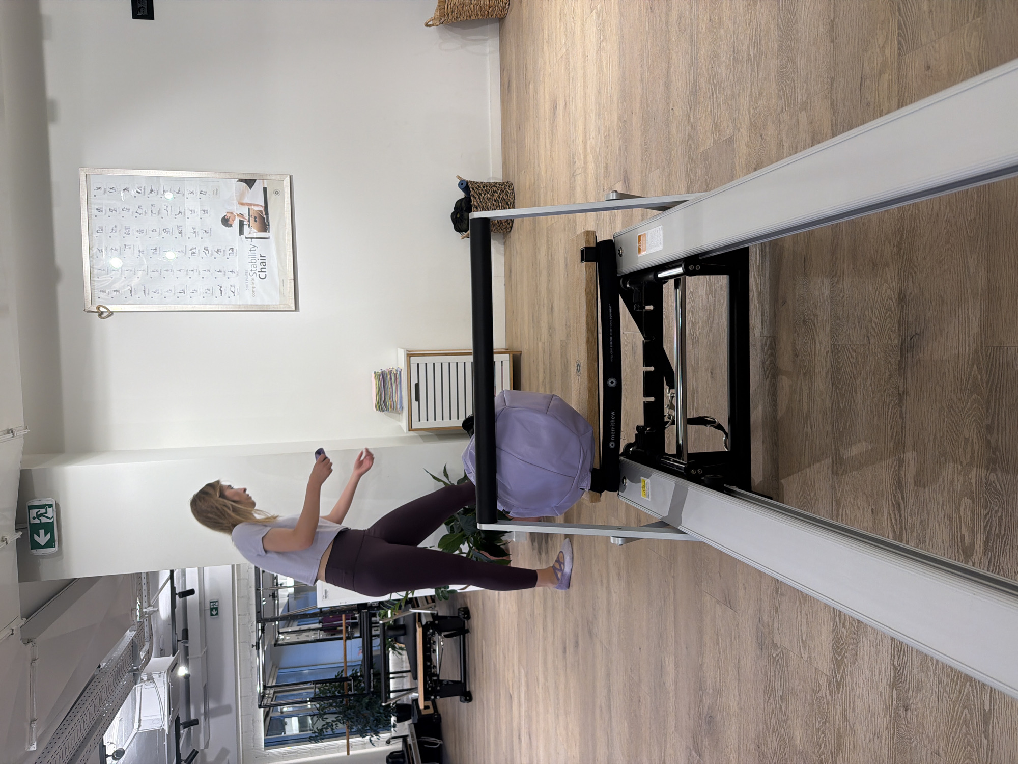 The image shows a blonde woman in a purple athletic outfit standing in a gym or studio. She is holding a phone and appears to be in the process of putting it away. In the foreground, a Pilates reformer machine is visible. The room has light wooden floors and white walls, with various gym equipment and a poster on the wall. The scene appears to be a fitness studio. There is no information in the image that would indicate its location in Boreti, Montenegro.