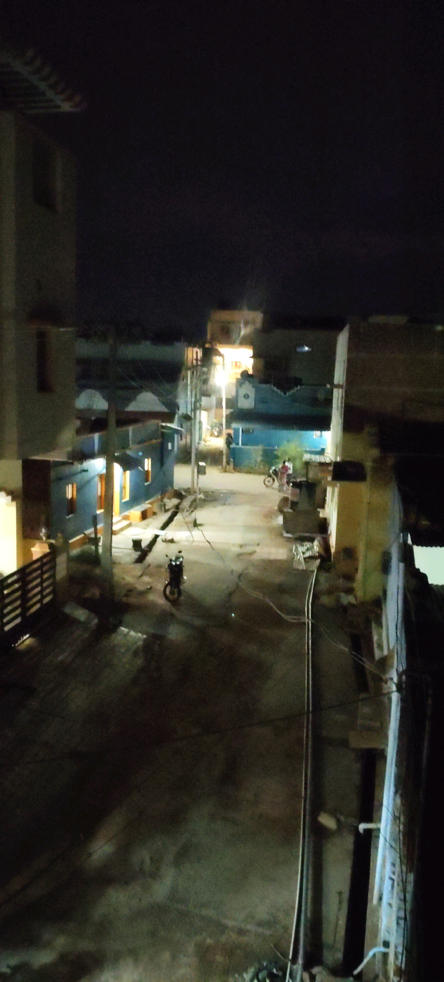 The image depicts a street scene at night in a residential area, likely in Erasakkanayakkanur, India, given the context. The perspective is from an elevated viewpoint, looking down onto a narrow street lined with buildings.
In the foreground, a dark, weathered street surface is visible, marked by shadows. To the left, a dark metal gate or fence runs along the edge of a property, with a dimly lit structure behind it. A dark-colored motorcycle is parked in the middle of the street. Further down the street, another motorcycle, also dark, is parked.
The buildings on either side of the street are multi-story structures, typical of Indian residential architecture. Some have brightly lit windows, suggesting occupancy, while others appear darker. The building on the left, closer to the viewer, has a distinctly modern, light-colored facade. Across the street, there are buildings with blue and yellow paint, some with visible windows illuminated from within.
In the mid-ground, two figures appear to be standing on the street near a building. One is on a motorcycle and the other is standing. The lighting is dim, with a streetlamp casting light on the scene and contributing to the shadows. The sky above is dark, indicating nighttime. Utility poles and wires are visible, traversing the street.
The overall impression is of a quiet, dimly lit urban or semi-urban environment after dark. The scene appears to be a typical residential street, with signs of daily life such as parked vehicles and illuminated homes.