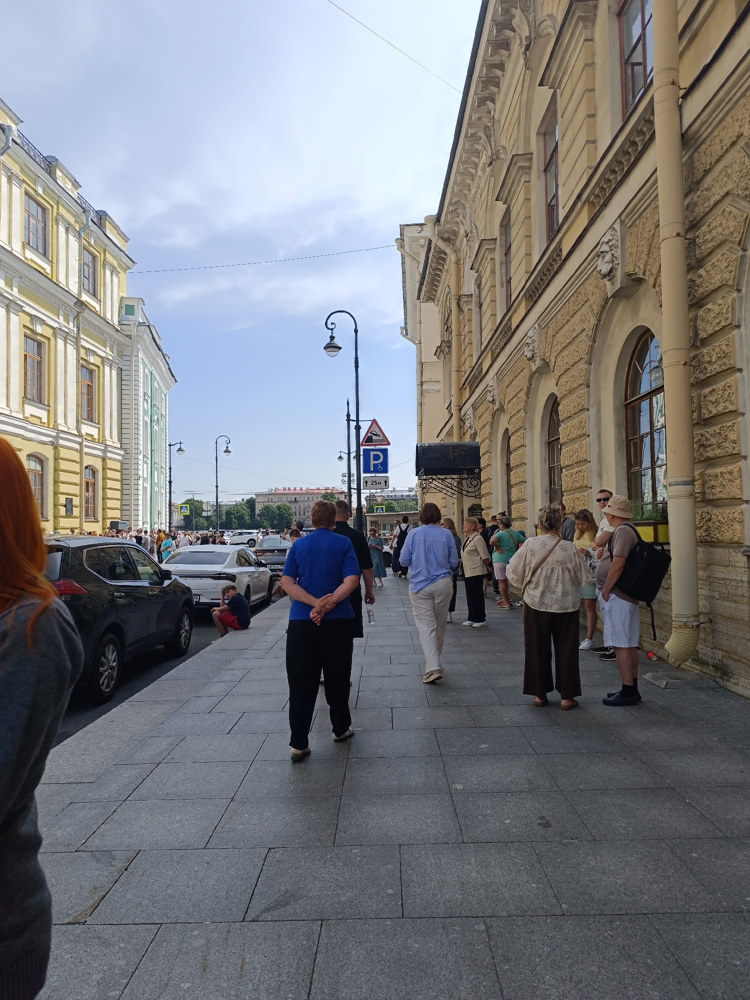 The image shows a street scene in Saint Petersburg, Russia, on a sunny day with a partly cloudy sky. A line of people is gathered on the sidewalk in front of an ornate, yellow building with classical architectural details, including columns and arched windows. The building appears to be a historical landmark. To the left, another yellow building with white trim and similar window styles lines the street. Cars are parked along the curb on the left side of the street, and a few pedestrians are walking on the sidewalk. A street lamp with an elaborate design stands near the center of the image, and a blue parking sign with a 'P' symbol is visible on the right side. The pavement is made of large, grey stone slabs. The overall impression is of a busy urban environment with a strong historical presence.