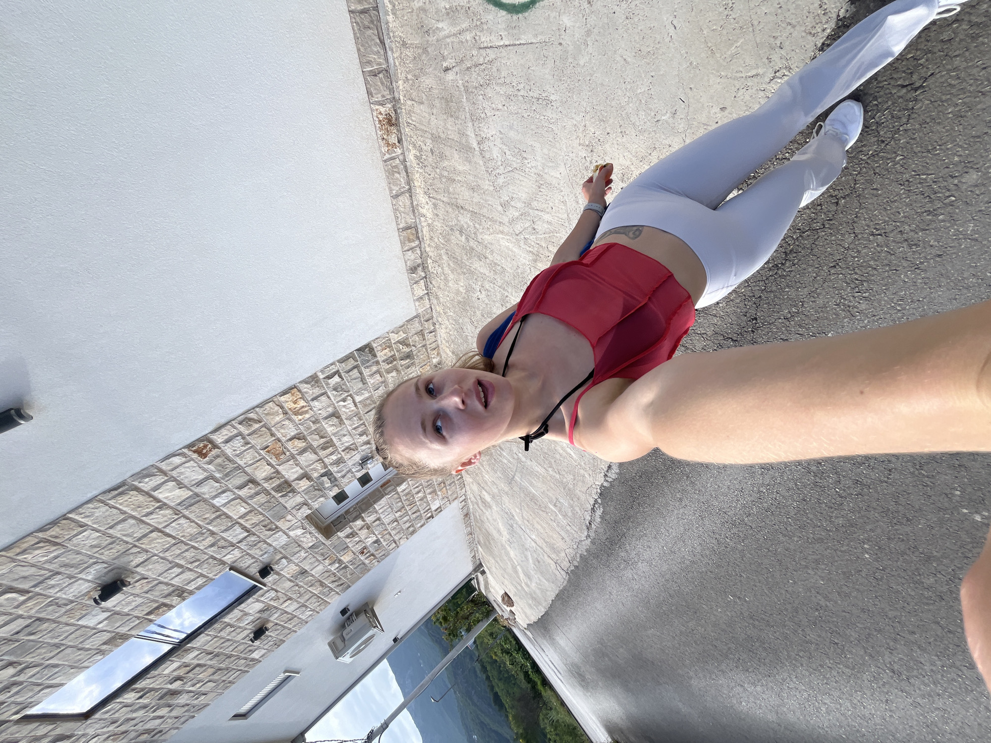 A female stands on an asphalt road in what appears to be Ivanovići, Montenegro. She wears white leggings, a red tank top, and white sneakers. A building with stone facade is visible on the left side of the frame, along with a distant mountain view in the background. The image appears to be a selfie.