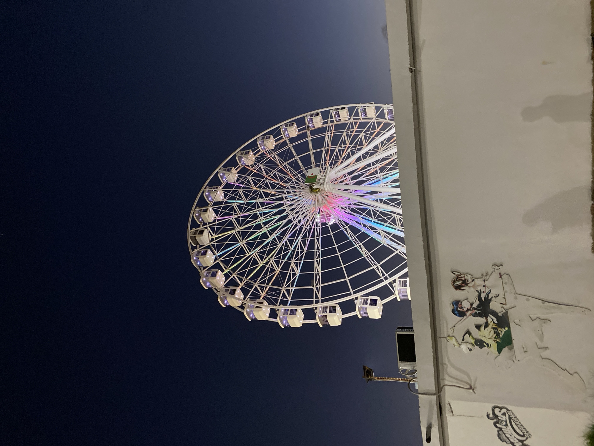 An illuminated Ferris wheel stands against a dark blue twilight sky. The Ferris wheel is white with multiple cabins, each lit with a purplish glow. The spokes of the wheel are outlined with colorful LED lights in pink, blue, and green hues. Below the Ferris wheel, a plain white concrete wall spans the lower portion of the image. On the left side of the wall, graffiti depicting cartoon characters can be seen. To the left of the graffiti, a street light pole extends upwards. The scene appears to be outdoors, possibly in an amusement park or public area within Hussein Dey, Algeria.