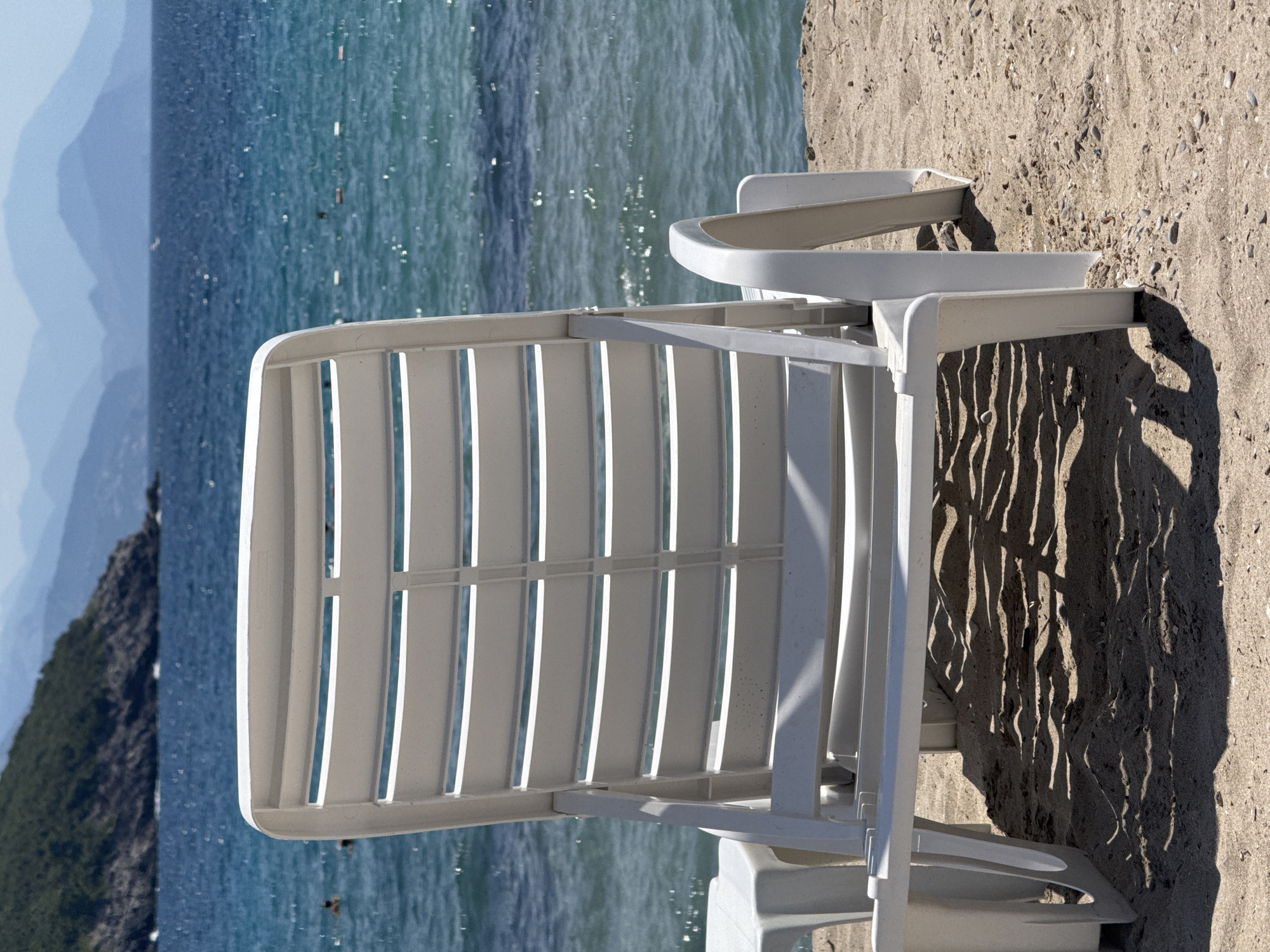 A white plastic beach chair faces the ocean from a sandy beach. The chair is in the foreground, with its back to the viewer, showing a slatted design. To the left of the chair is a portion of a matching white plastic side table. The sand is textured with shadows cast from the chair and table, suggesting a sunny day. In the mid-ground, the turquoise ocean stretches out, with a few people visible swimming or wading in the water. Further in the distance, a green, tree-covered hillside juts out into the sea on the left side of the frame. Beyond that, layers of hazy blue mountains recede into the horizon under a bright sky.