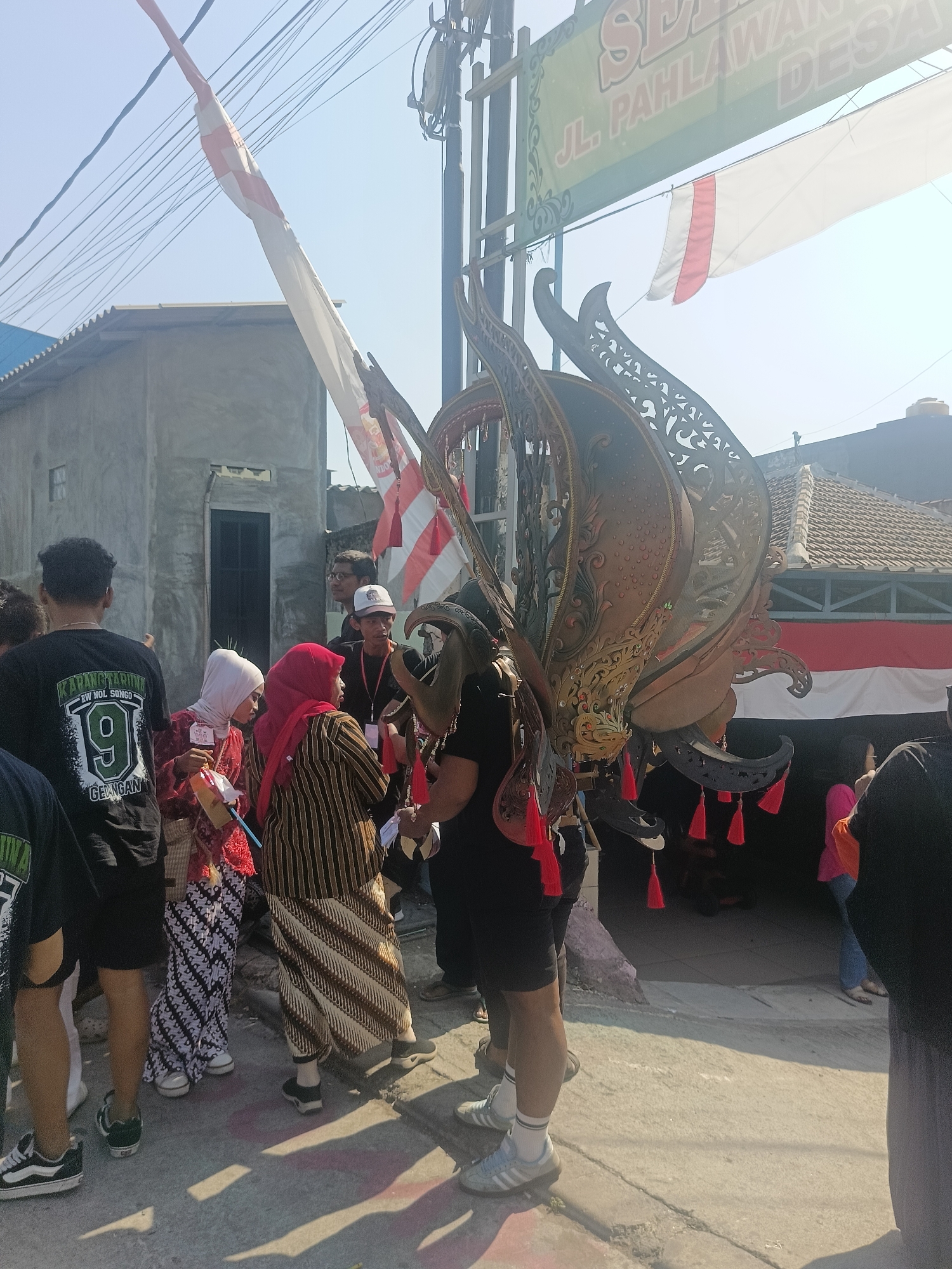 This image captures a scene outdoors, likely in Gedangan, Indonesia, with people gathered. A prominent feature is a large, ornate decorative headdress or costume piece, resembling a bird's wings and head, adorned with intricate carvings and red tassels. Several individuals are in the foreground, some wearing traditional Indonesian attire, such as batik patterns. One person in the center wears a red hijab and a striped, long-sleeved top with a batik skirt. Another person to their left wears a white hijab and a red patterned garment. A man in the background wears a black t-shirt with a graphic that reads "KARANG TARUNA RW NOL SONGO 9 GELINGAN". There are also Indonesian flags, one red and white, and another white flag with red stripes visible in the background, suggesting a festive or commemorative event. Buildings with concrete walls and tiled roofs line the street, and power lines are strung overhead. The ground appears to be a paved surface, possibly a sidewalk or road, with shadows cast by the sunlight.