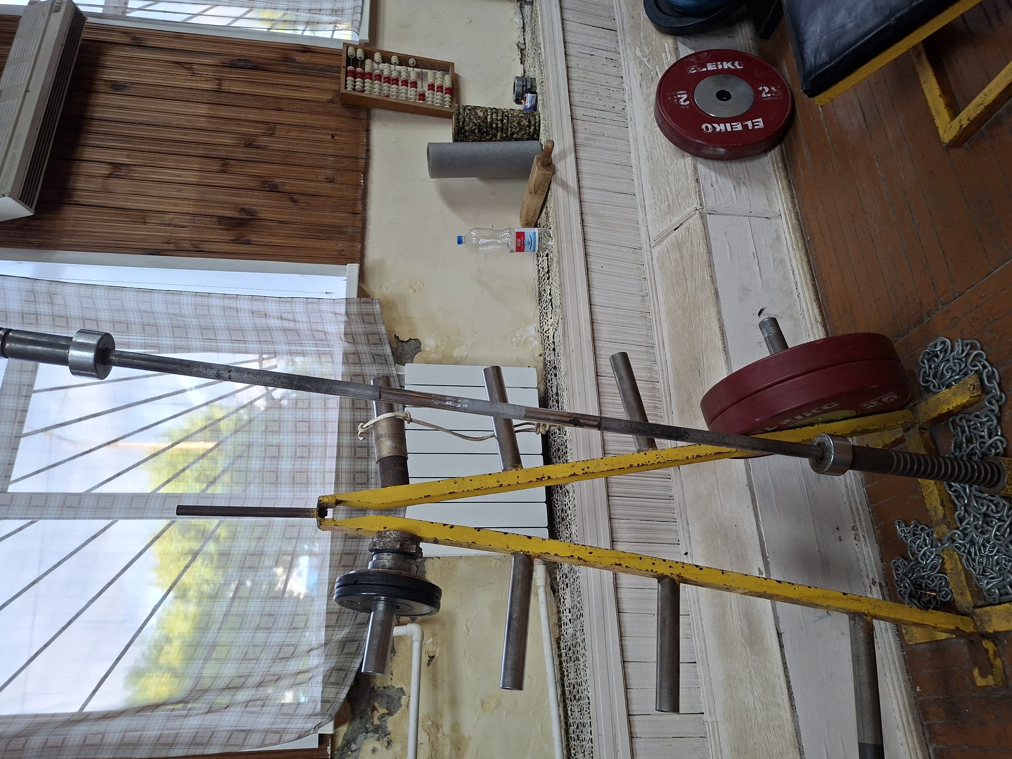 This image depicts an indoor gym setting, possibly in Kazan, Russia, with various weightlifting equipment. In the foreground, a yellow weight rack holds a barbell with weight plates. Chains are coiled at the base of the rack. To the right, a red weight plate labeled "ELEIKO 25" rests on the floor. Further to the right, a black and yellow weight bench is partially visible.
The background reveals a window covered by a checkered curtain, with trees visible through the glass. The walls are painted a light, textured color, with some areas showing signs of wear. A wooden paneling runs along the upper portion of the wall. On a shelf above the bench, a plastic water bottle, two foam rollers (one grey, one patterned), and a wooden rolling pin are present. To the far right, an abacus-like device with red and white beads is mounted on the wall. The floor is made of worn wooden planks. No people are present in the image.