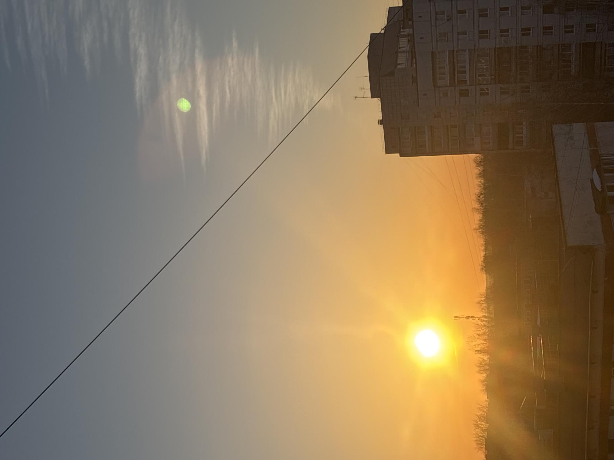 The image depicts a sunset over a cityscape, likely in Chaikovsky, Russia. The sun is prominent in the lower right, casting a golden glow over the scene. A tall building with many windows stands to the right of the sun, silhouetted against the bright light. A wire stretches diagonally across the sky from the upper right to the lower left. The sky transitions from a pale blue in the upper left to a warm orange near the sun. Wispy clouds are visible in the blue portion of the sky. Bare trees are visible along the horizon line near the sun. The angle of the photograph suggests it was taken from a high vantage point.