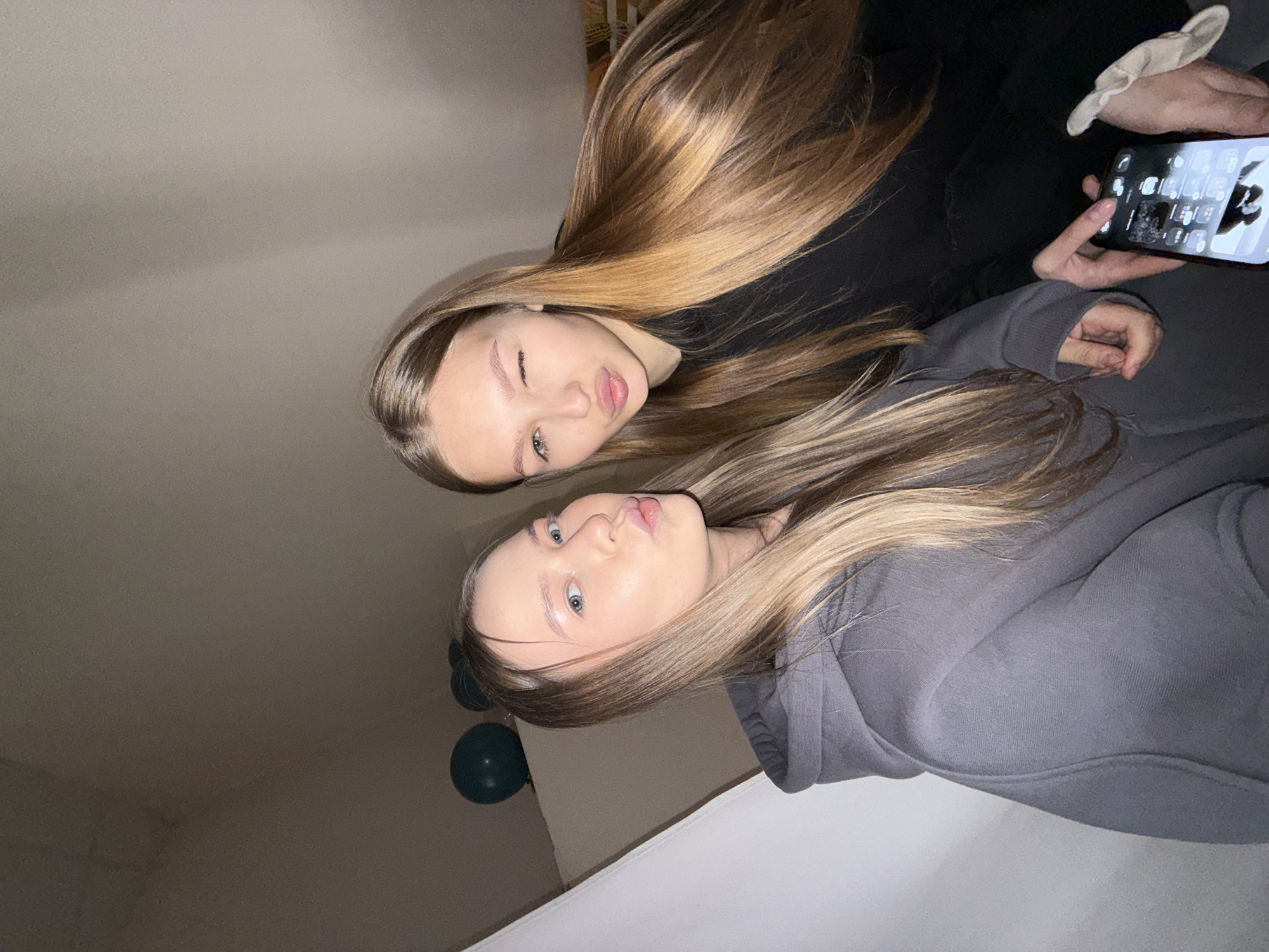 Two young women are captured in a close-up, upward-angle shot, likely a selfie, indoors. The person in the upper part of the frame has long, straight, light brown hair with blonde highlights, and is wearing a black top or hoodie. She is winking with one eye and slightly puckering her lips, looking directly at the camera. She holds a smartphone in her right hand, its screen showing a grid of app icons. Her fingernails have a light-colored polish.
The second person is positioned below and to the right, also with long, straight, light brown or blonde hair, and is wearing a grey hoodie. She looks towards the camera with slightly puckered lips. Both individuals have fair complexions and light-colored eyes.
The background is a plain, light-colored wall and ceiling, suggesting a room. Two dark-colored, possibly black or dark blue, balloons are partially visible in the lower left corner of the frame, hinting at a casual event or celebration. The lighting is artificial and somewhat dim, casting soft shadows. The overall tone of the image is casual and playful. No distinct text is legible other than the blurry app icons on the phone screen. The location context provided is Chaikovsky, Russia, though no specific visual elements in the image directly confirm this.