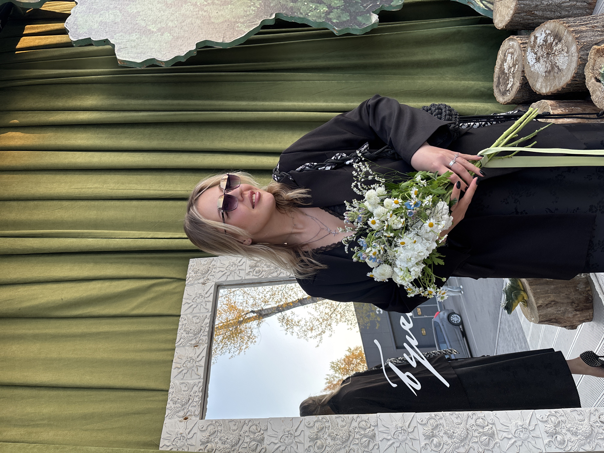 A woman is holding a bouquet of flowers in front of a mirror. She is wearing sunglasses, a black dress with a blazer, and has blonde hair. Behind her is a green curtain and some cut logs. In the reflection of the mirror, you can see a tree and a car. The location is Krasnoyarsk, Russia.