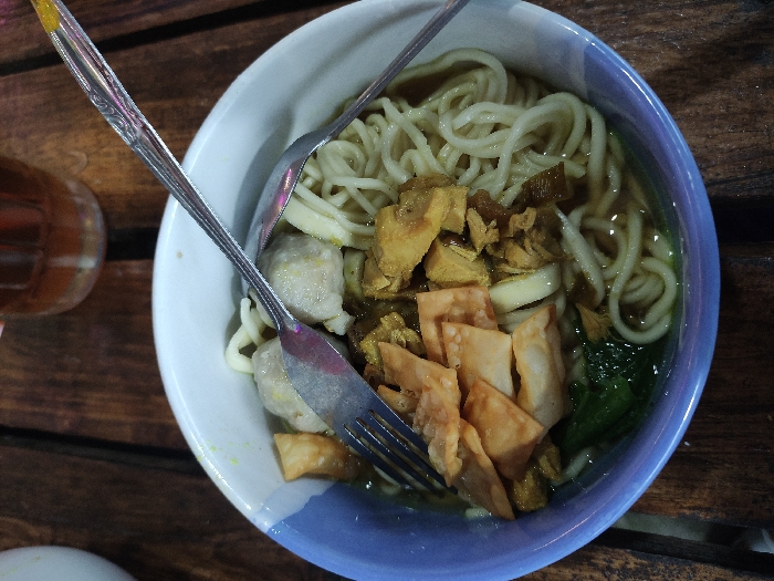 An overhead shot captures a bowl of Indonesian noodles, likely Mie Ayam or Mie Bakso, placed on a dark wooden table in Kecamatan Pandaan, Indonesia. The round bowl features a white interior and a light blue rim. Inside, a generous serving of thin, pale noodles rests in a light brown broth.
Accompanying the noodles are various toppings: chunks of what appears to be shredded or diced chicken, several pale, round meatballs (bakso), pieces of green leafy vegetables (possibly bok choy or mustard greens), and golden-brown, crispy fried wonton skins. A metal fork and spoon are partially submerged in the bowl, suggesting the meal is either about to be eaten or is in progress.
To the left of the bowl, a portion of a glass containing a amber-colored beverage, possibly iced tea, is visible. The wooden table has a rustic appearance with visible grain and gaps between the planks. The lighting is dim, indicating an indoor setting or an evening/nighttime meal. No text is visible in the image.