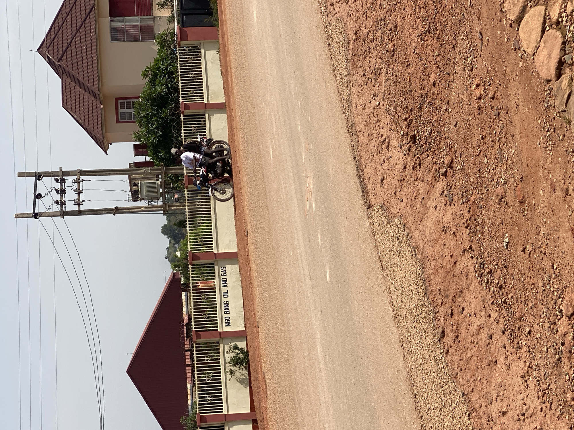 The image captures a street scene in Jos, Nigeria, under a bright, clear sky. In the foreground, a reddish-brown, unpaved shoulder borders a paved road. Beyond the road, a white fence with red posts and decorative heart shapes encloses a property. A sign on the wall reads "NGO BANG OIL AND GAS." To the left of the sign, a dark red, gabled roof building is visible. To the right, a light-colored house with a brown tiled roof stands, featuring red-trimmed windows and a cross on its peak. A large electrical transformer and utility pole are situated behind the fence, near the house. On the road, two individuals are riding a black motorcycle. One person, wearing a white shirt and a gray beanie, is driving, while the other, wearing a dark shirt, is the passenger. They are moving away from the viewer. Lush green trees and bushes are interspersed throughout the scene, adding to the outdoor setting.