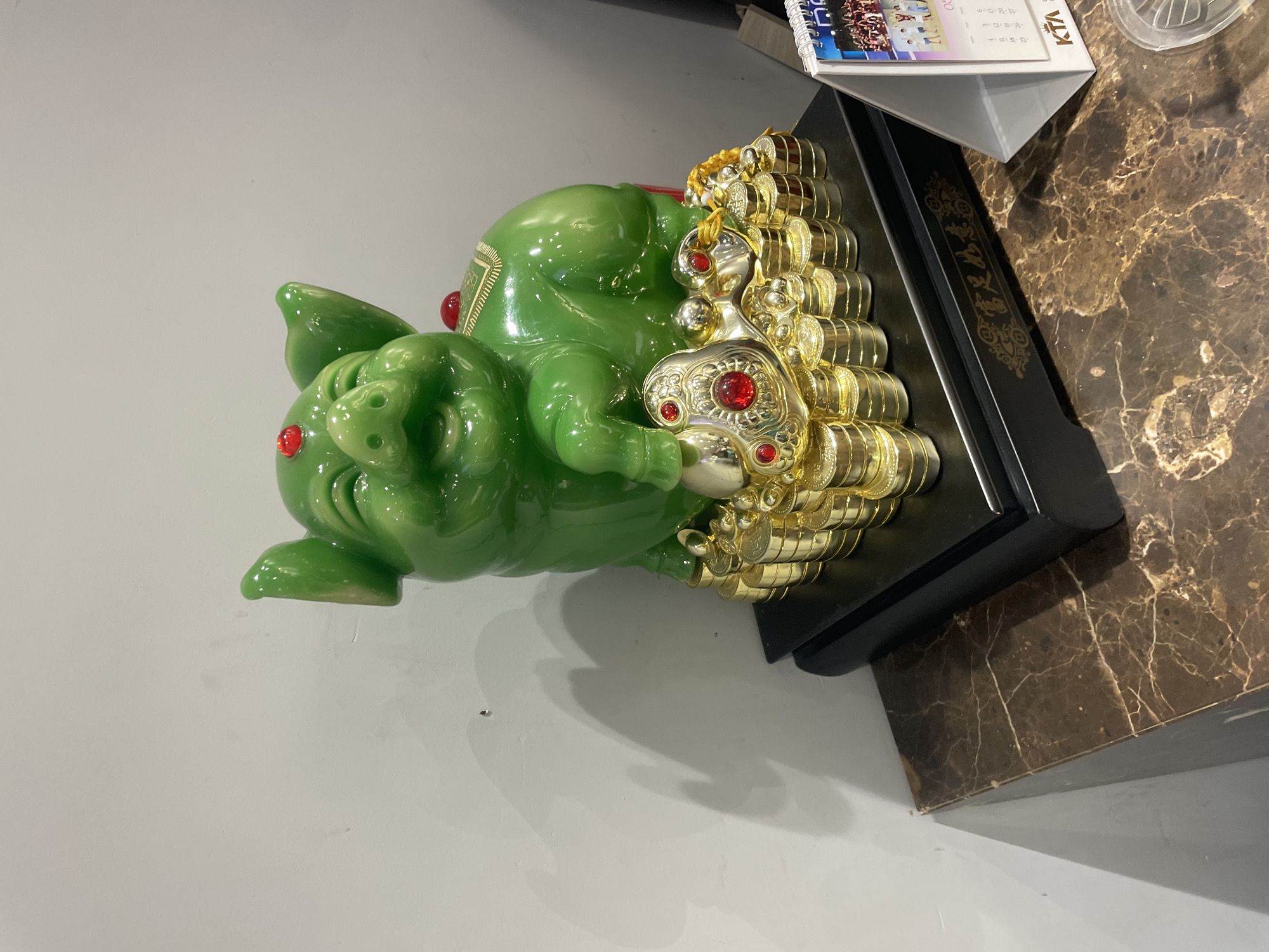 A glossy green pig figurine sits on a stack of gold coins, which are on a black base. The figurine has red jewel accents on its forehead and back, and a large gold and red jewel ornament in front of it. The base has gold Chinese characters and designs. The pig is on a brown marble-patterned surface, next to a white desk calendar. The background is a plain light grey wall with shadows cast by the figurine. This scene is located in Thủ Đức City, Vietnam.