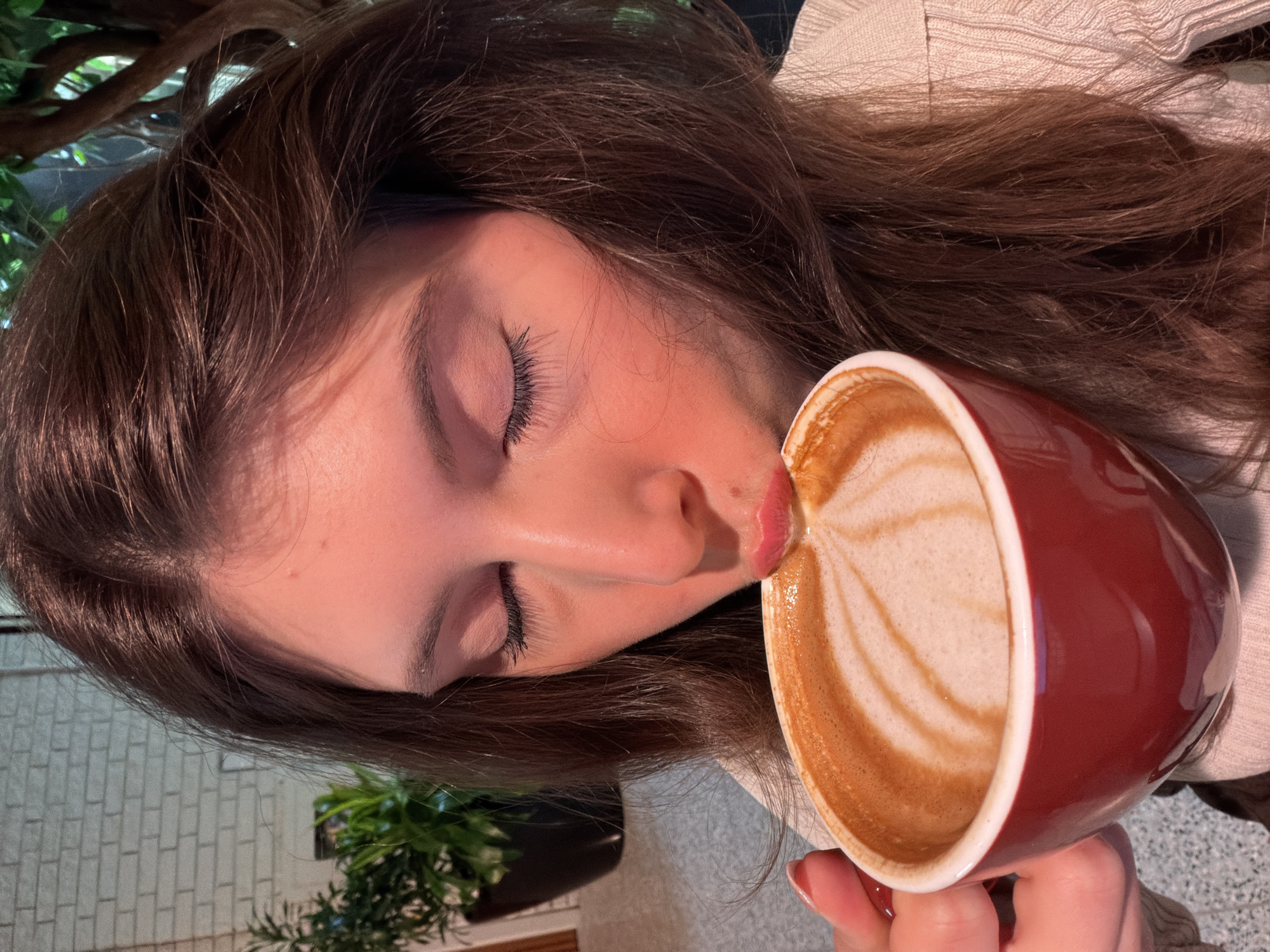 A woman with long brown hair is holding a red mug containing a latte with latte art. Her eyes are closed, and her lips are touching the rim of the mug as if she is about to take a sip. The scene appears to be indoors, possibly in a cafe or restaurant, based on the presence of plants and what might be a wooden beam in the background. The location is not discernible from the image, so it's impossible to confirm if it is in Montreal, Canada.