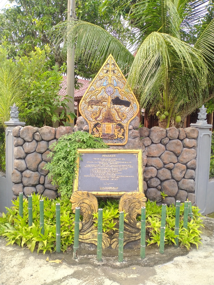 This daytime outdoor shot captures a monument in Karanganyar, Indonesia, set against a backdrop of lush green foliage and a clear sky. The central feature is a stele-like structure, part of a stone-patterned wall. Atop this wall stands a prominent, intricately carved golden "gunungan" (a traditional Javanese wayang kulit motif representing a mountain or tree of life). The gunungan depicts symbolic figures and patterns, including what appear to be a house/temple structure, animal forms (possibly a tiger), and birds. Below the gunungan, a rectangular black plaque framed in gold is mounted on the wall. The base of the monument is surrounded by vibrant green plants, with a semi-circle of short, green bamboo poles arranged in front of it, acting as a decorative boundary. In the background, tall green trees, including palm trees laden with coconuts, are visible. A building with a red-tiled roof is partially obscured by the dense vegetation on the right. The lighting suggests a clear and bright day. The visible text on the plaque reads: "PRASASTI" (Inscription/Charter) "Visi Kementerian Agraria dan Tata Ruang/Badan Pertanahan Nasional: TERWUJUDNYA PENATAAU RUANG DAN PERTANAN YANO BERKEADILAN DAN KEBERLANJUTAN UNTUK KEMAKMURAN RAKYAT" (Vision of the Ministry of Agrarian Affairs and Spatial Planning/National Land Agency: The realization of fair and sustainable spatial planning and land management for the prosperity of the people) "VISI PEMBANGUNAN KABUPATEN KARANGANYAR 2018-2023: MAJU DAN INOVATIF DENGAN DIDUKUNG OLEH MASYARAKAT YANG MANDIRI, SEJAHTERA, DAN BERKARAKTER" (Vision for the Development of Karanganyar Regency 2018-2023: Advanced and Innovative, supported by an independent, prosperous, and characterful society) "Pesan:" (Message:) 1. "Peran Pertanahan adalah mengembalikan Hak Rakyat, sebagai Pondasi Kehidupan Masyarakat, untuk dapat hidup layak" (The role of Land Affairs is to restore the rights of the people, as the foundation of community life, to live decently) 2. "Pertanahan sebagai Pondasi, dalam meningkatkan Harkat dan Martabat Rakyat" (Land Affairs as a foundation, in improving the dignity and honor of the people) 3. "Pertanahan sebagai pondasi, membangun Ekonomi Rakyat" (Land Affairs as a foundation, building the people's economy) 4. "Pertanahan sebagai pondasi, dalam menciptakan Kedamaian" (Land Affairs as a foundation, in creating peace) 5. "Pertanahan sebagai Pondasi, mewujudkan Kebahagiaan Rakyat" (Land Affairs as a foundation, realizing the happiness of the people) Dated "YOGYAKARTA, ... JANUARI 2018" Signed "A.n. MENTERI AGRARIA DAN TATA RUANG/ KEPALA BADAN PERTANAHAN NASIONAL DR. Ir. H. SUDIJONO MS. DIREKTUR JENDERAL HUBUNGAN HUKUM PERTANAHAN" (On behalf of the Minister of Agrarian
